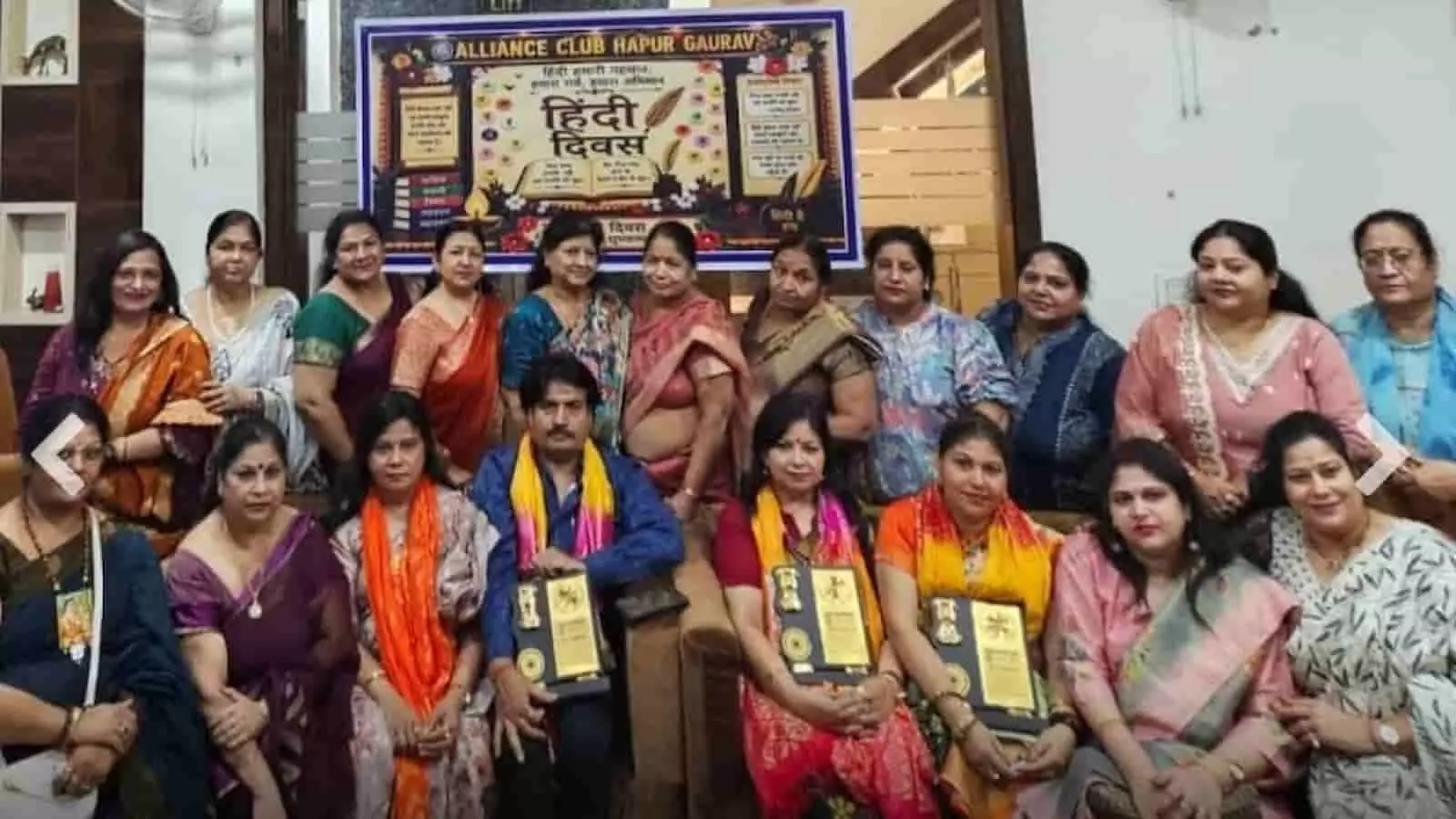 तस्वीर में बाएं से दाएं बैठी और खड़ी महिलाओं तथा एक पुरुष के समूह के पीछे हिंदी दिवस का बैनर लगा हुआ है। तस्वीर में बाएं से दाएं बैठी और खड़ी महिलाओं तथा एक पुरुष के समूह के पीछे हिंदी दिवस का बैनर लगा हुआ है।