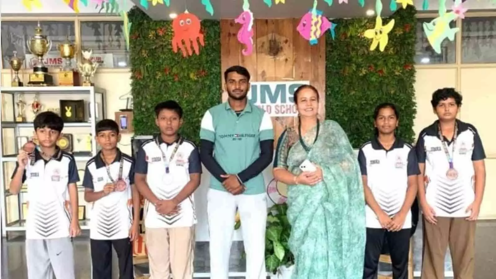 तस्वीर में बाएं से दाएं चार छात्र, एक कोच, एक शिक्षिका और दो अन्य छात्र पदक पहने हुए खड़े हैं। तस्वीर में बाएं से दाएं चार छात्र, एक कोच, एक शिक्षिका और दो अन्य छात्र पदक पहने हुए खड़े हैं।