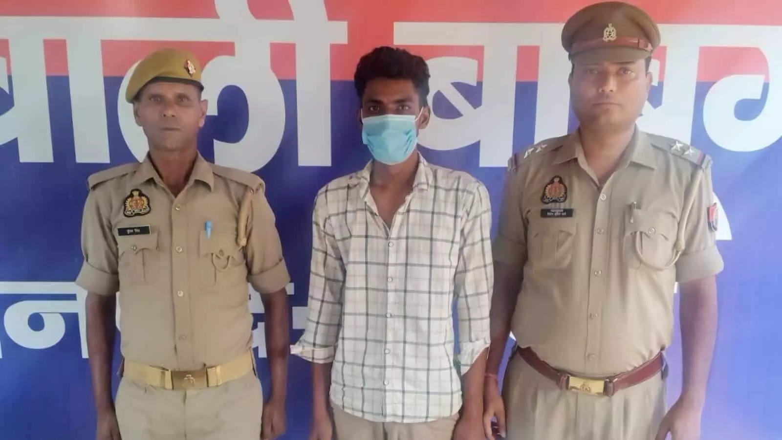 तस्वीर में दो पुलिसकर्मियों के बीच खड़े चेकदार शर्ट और मास्क पहने आरोपी के पीछे उत्तर प्रदेश पुलिस का बोर्ड नजर आ रहा है। तस्वीर में दो पुलिसकर्मियों के बीच खड़े चेकदार शर्ट और मास्क पहने आरोपी के पीछे उत्तर प्रदेश पुलिस का बोर्ड नजर आ रहा है।