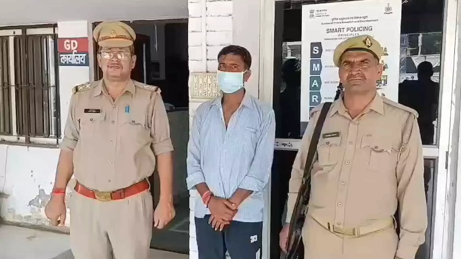 तस्वीर में बाईं और दाईं ओर दो पुलिस अधिकारियों के बीच खड़े नीली धारीदार शर्ट और मास्क पहने आरोपी के पीछे एक कार्यालय का दरवाजा नजर आ रहा है। तस्वीर में बाईं और दाईं ओर दो पुलिस अधिकारियों के बीच खड़े नीली धारीदार शर्ट और मास्क पहने आरोपी के पीछे एक कार्यालय का दरवाजा नजर आ रहा है।