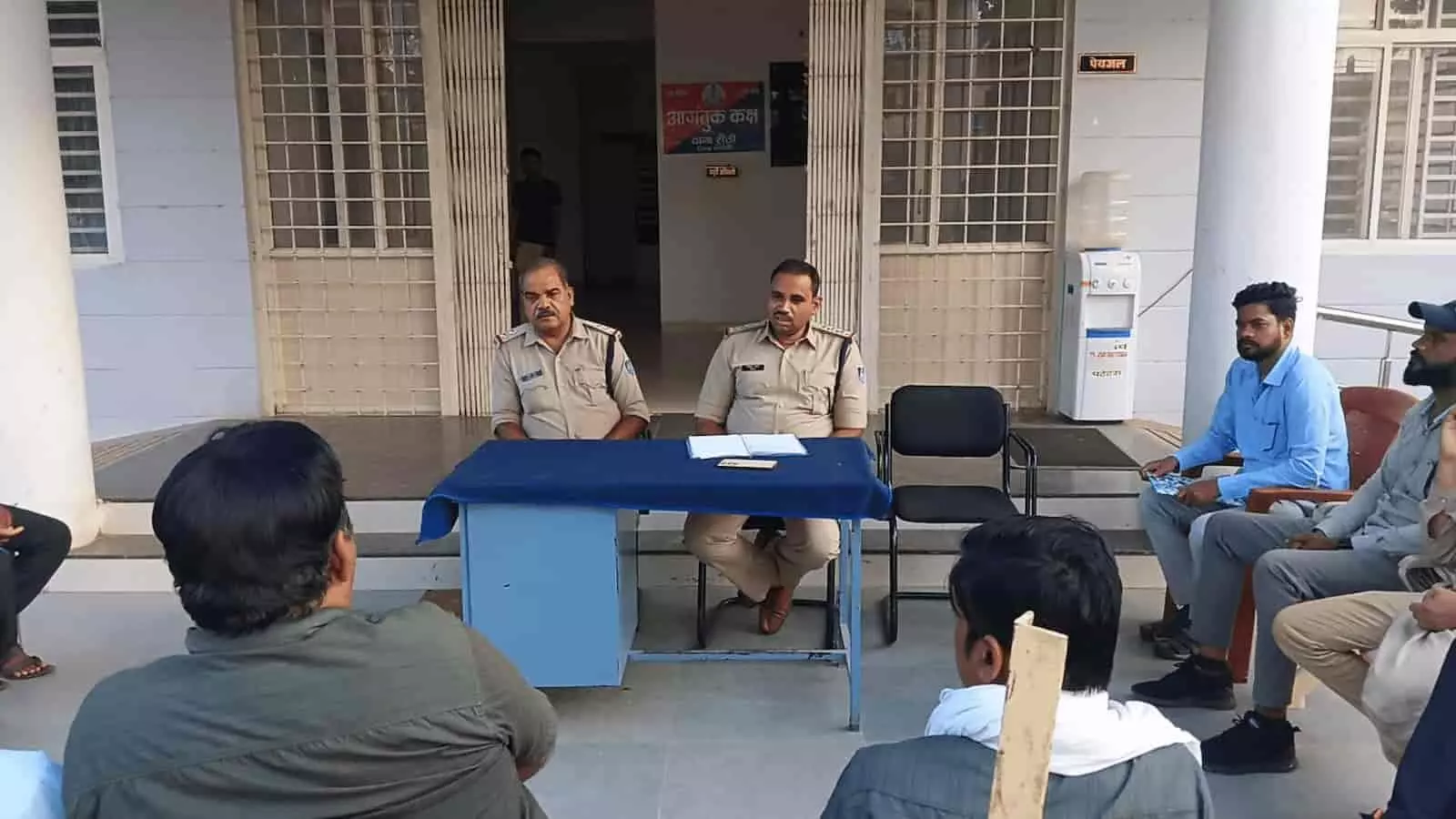 तस्वीर में नीले कपड़े से ढकी मेज के पीछे बैठे दो पुलिस अधिकारियों के साथ आसपास नागरिक कुर्सियों पर बैठे दिखाई दे रहे हैं। तस्वीर में नीले कपड़े से ढकी मेज के पीछे बैठे दो पुलिस अधिकारियों के साथ आसपास नागरिक कुर्सियों पर बैठे दिखाई दे रहे हैं।