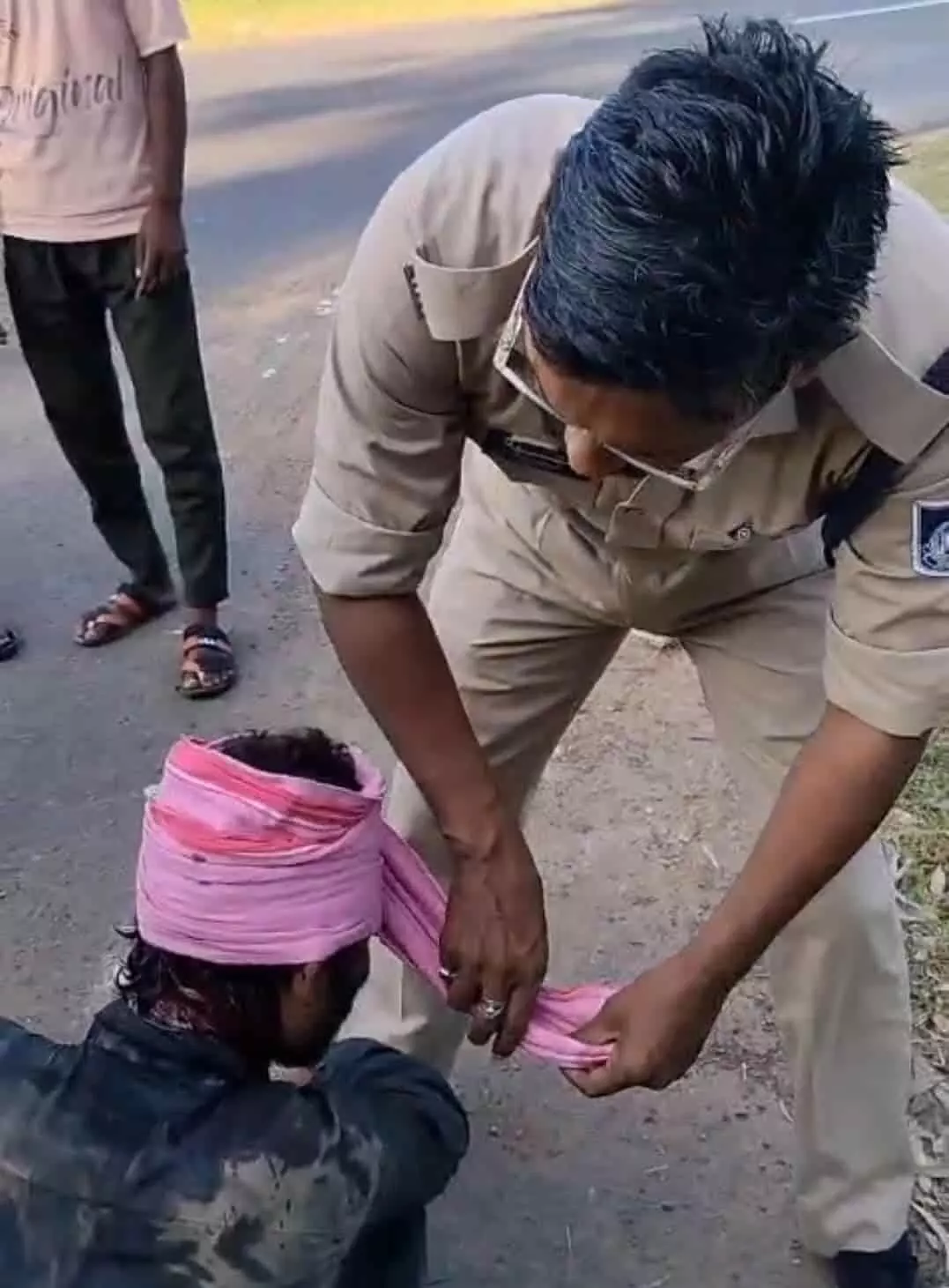 तस्वीर में खाकी वर्दी पहने पुलिसकर्मी सड़क के किनारे बैठे घायल युवक के सिर पर गुलाबी गमछे से पट्टी बांध रहे हैं। तस्वीर में खाकी वर्दी पहने पुलिसकर्मी सड़क के किनारे बैठे घायल युवक के सिर पर गुलाबी गमछे से पट्टी बांध रहे हैं।