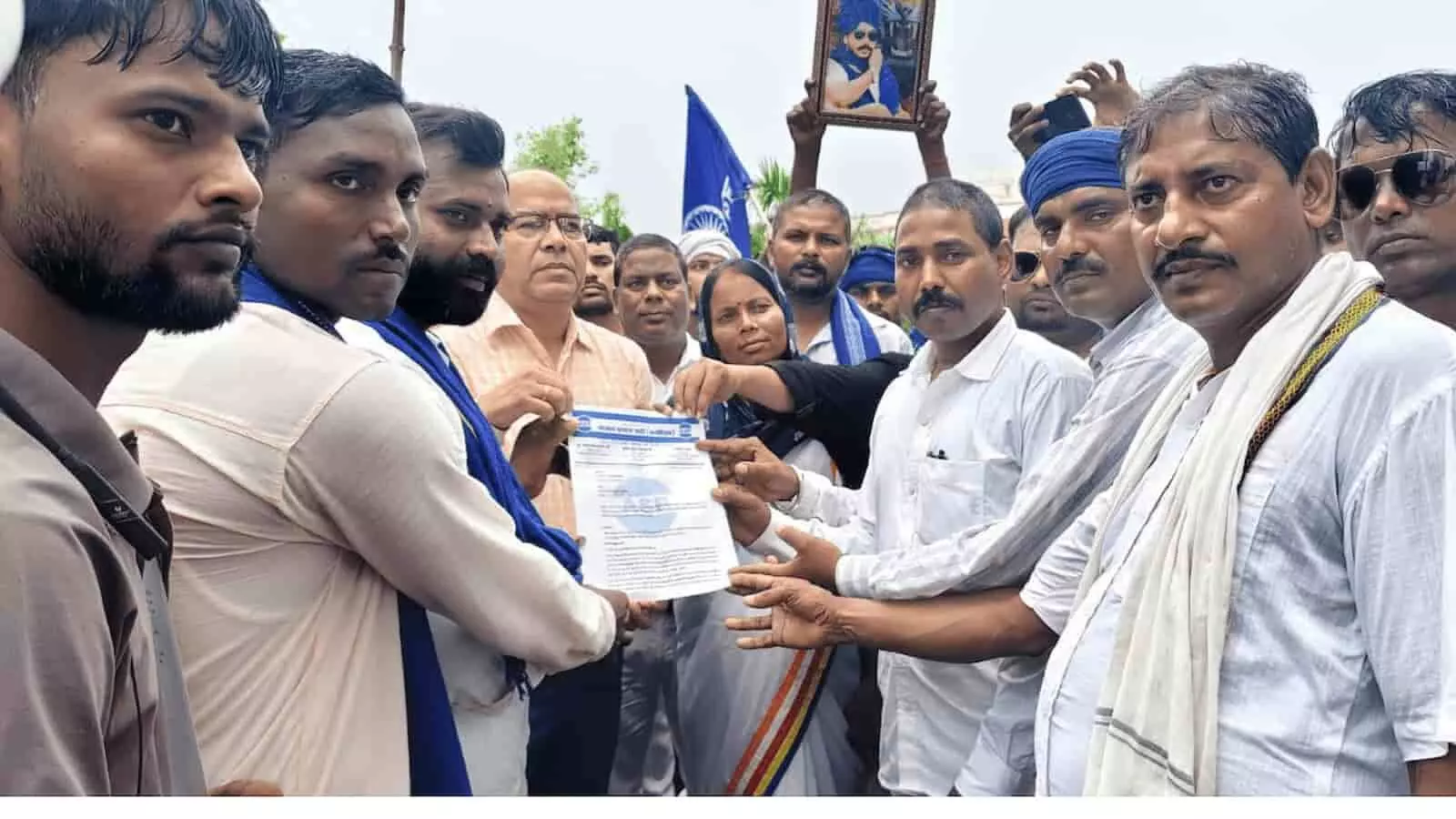 तस्वीर में कुछ लोग नीले झंडे और एक ज्ञापन को पकड़े हुए समूह में खड़े दिखाई दे रहे हैं। तस्वीर में कुछ लोग नीले झंडे और एक ज्ञापन को पकड़े हुए समूह में खड़े दिखाई दे रहे हैं।