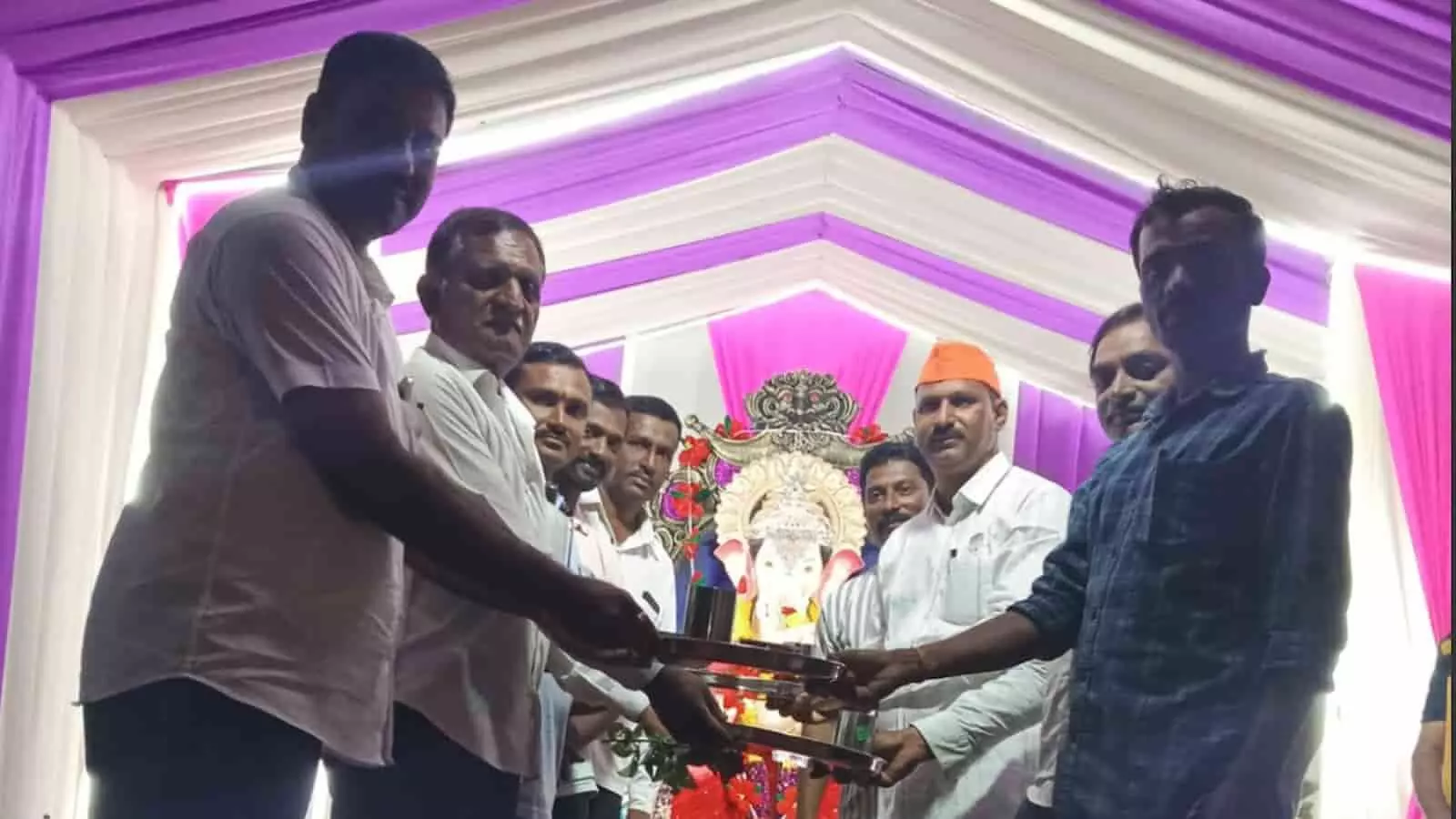 तस्वीर में पंडाल के भीतर भगवान गणेश की प्रतिमा के सामने खड़े कुछ लोग एक-दूसरे को स्टील की थालियां और बर्तन सौंपते हुए दिखाई दे रहे हैं. तस्वीर में पंडाल के भीतर भगवान गणेश की प्रतिमा के सामने खड़े कुछ लोग एक-दूसरे को स्टील की थालियां और बर्तन सौंपते हुए दिखाई दे रहे हैं.