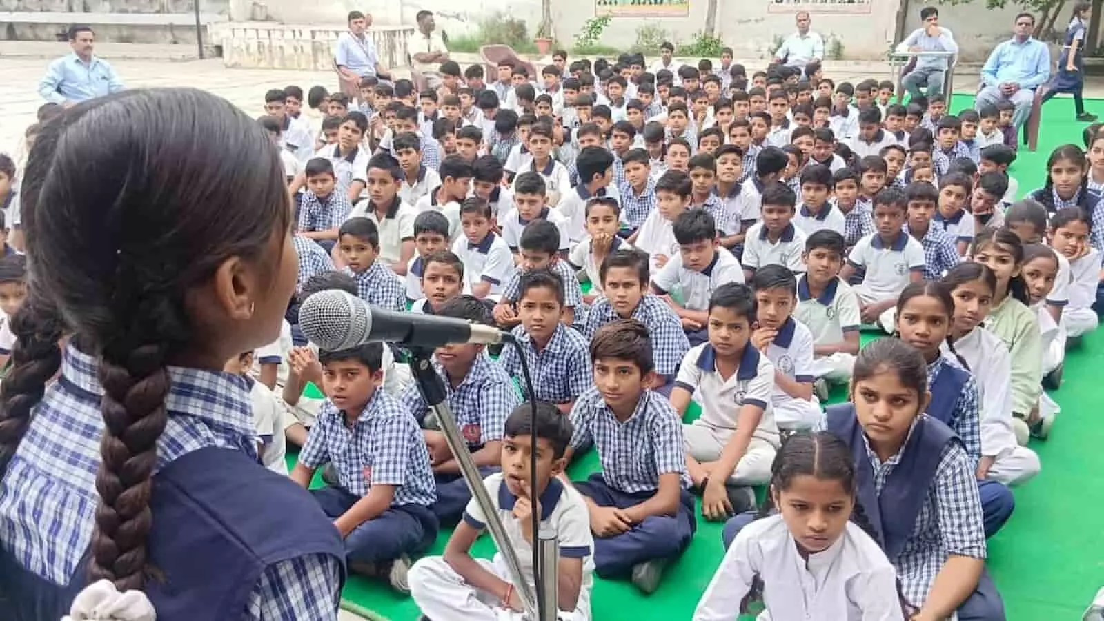 तस्वीर में स्कूल परिसर में जमीन पर बैठे विद्यार्थियों के समूह के सामने माइक पर कविता पाठ करती हुई एक छात्रा दिखाई दे रही है। तस्वीर में स्कूल परिसर में जमीन पर बैठे विद्यार्थियों के समूह के सामने माइक पर कविता पाठ करती हुई एक छात्रा दिखाई दे रही है।