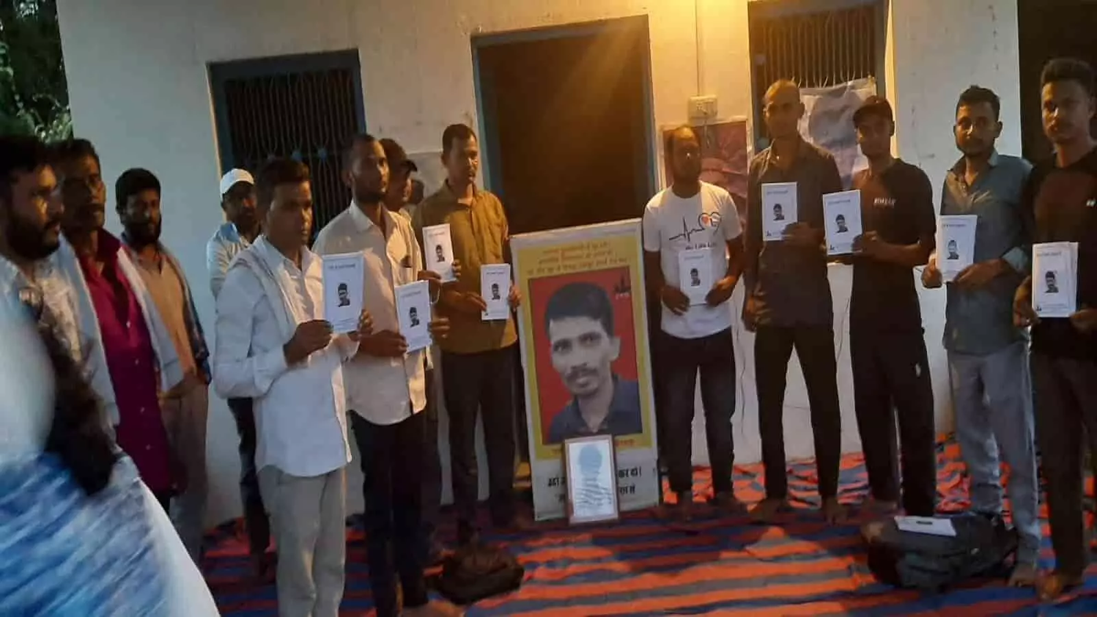 तस्वीर में दिख रही सभा में उपस्थित पुरुष एक बैनर के सामने हाथ में पुस्तिकाएं प्रदर्शित कर रहे हैं। तस्वीर में दिख रही सभा में उपस्थित पुरुष एक बैनर के सामने हाथ में पुस्तिकाएं प्रदर्शित कर रहे हैं।