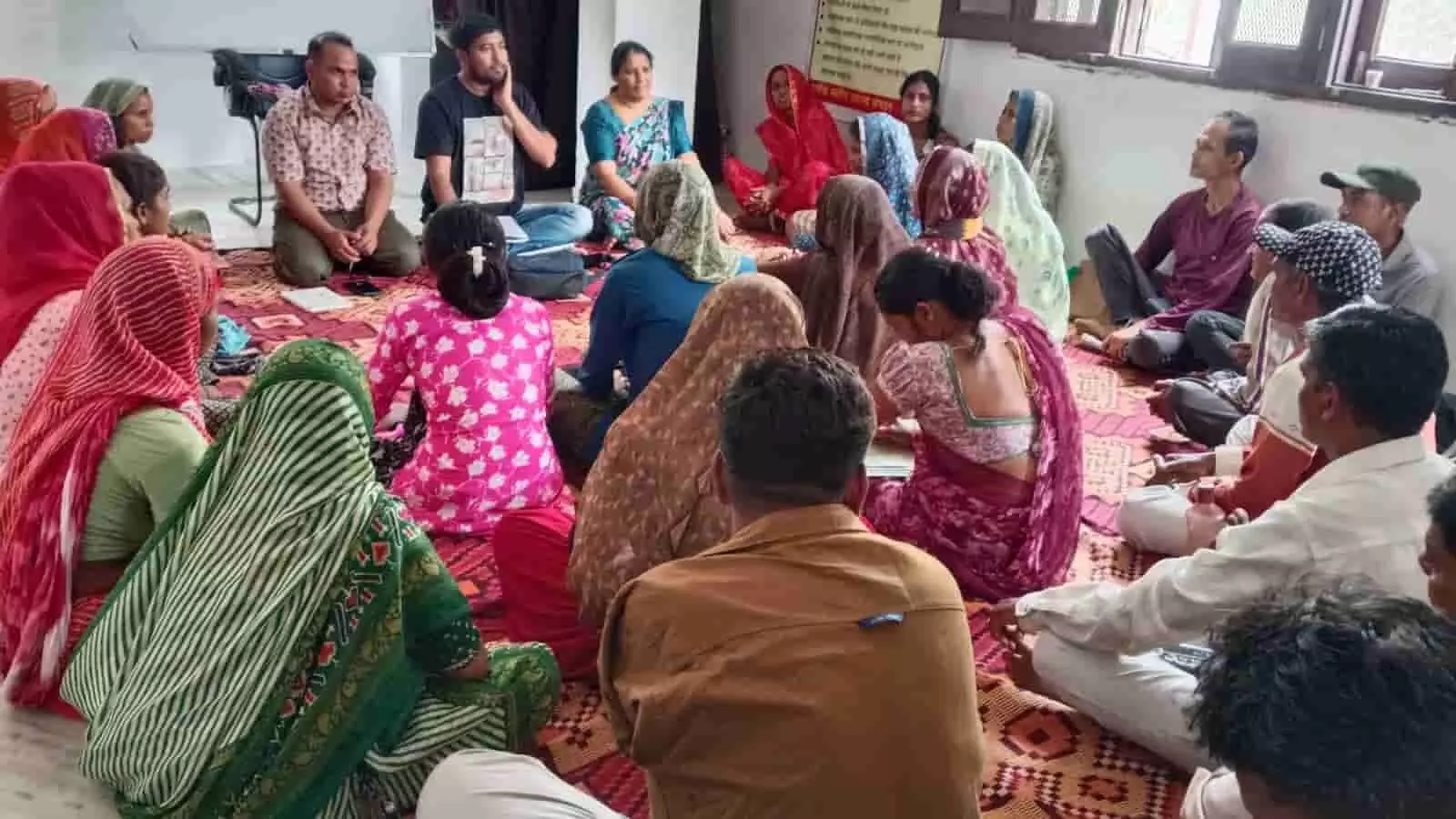 तस्वीर में दिख रही एक कमरे के भीतर जमीन पर बैठकर चर्चा करते पुरुष और महिलाएं। तस्वीर में दिख रही एक कमरे के भीतर जमीन पर बैठकर चर्चा करते पुरुष और महिलाएं।