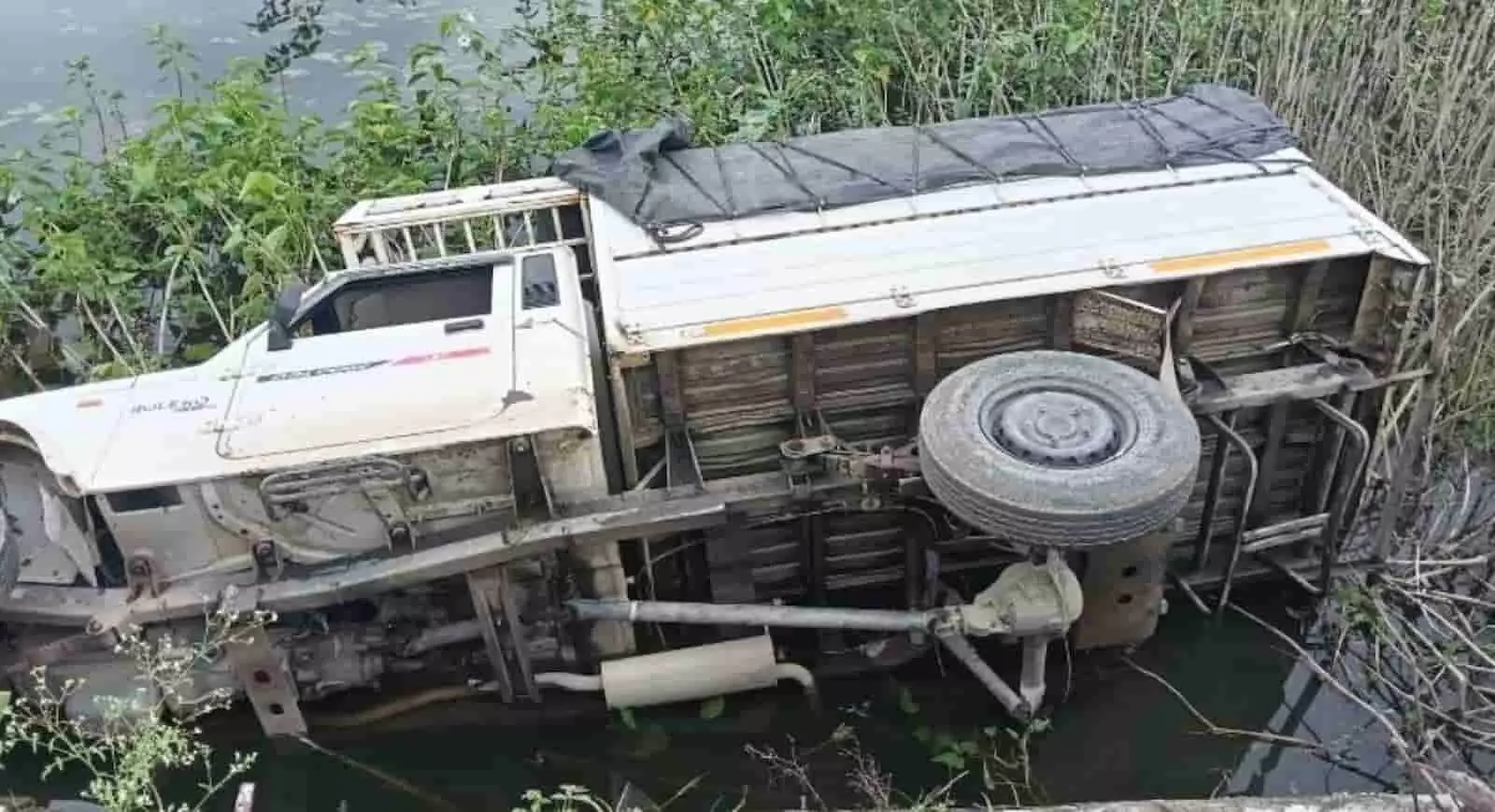 तस्वीर में एक सफेद पिकअप वाहन झाड़ियों और पानी के बीच करवट से पलटा दिख रहा है। तस्वीर में एक सफेद पिकअप वाहन झाड़ियों और पानी के बीच करवट से पलटा दिख रहा है।