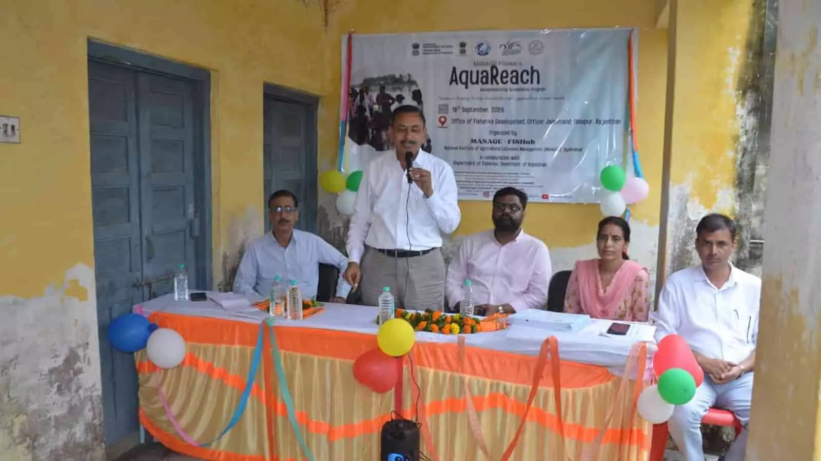 तस्वीर में दिख रही मेज के पीछे बैठे अधिकारियों के बीच एक व्यक्ति माइक पर बोल रहा है। तस्वीर में दिख रही मेज के पीछे बैठे अधिकारियों के बीच एक व्यक्ति माइक पर बोल रहा है।