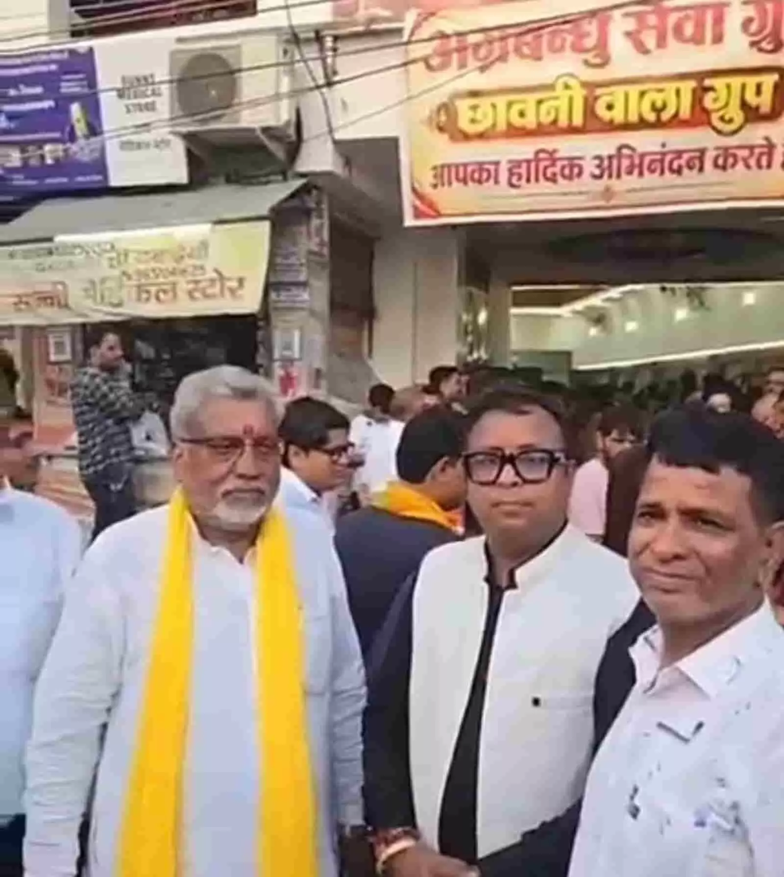 तस्वीर में दिख रही भीड़ के बीच में तीन पुरुष चुनावी कार्यालय के बैनर के आगे खड़े हैं। तस्वीर में दिख रही भीड़ के बीच में तीन पुरुष चुनावी कार्यालय के बैनर के आगे खड़े हैं।