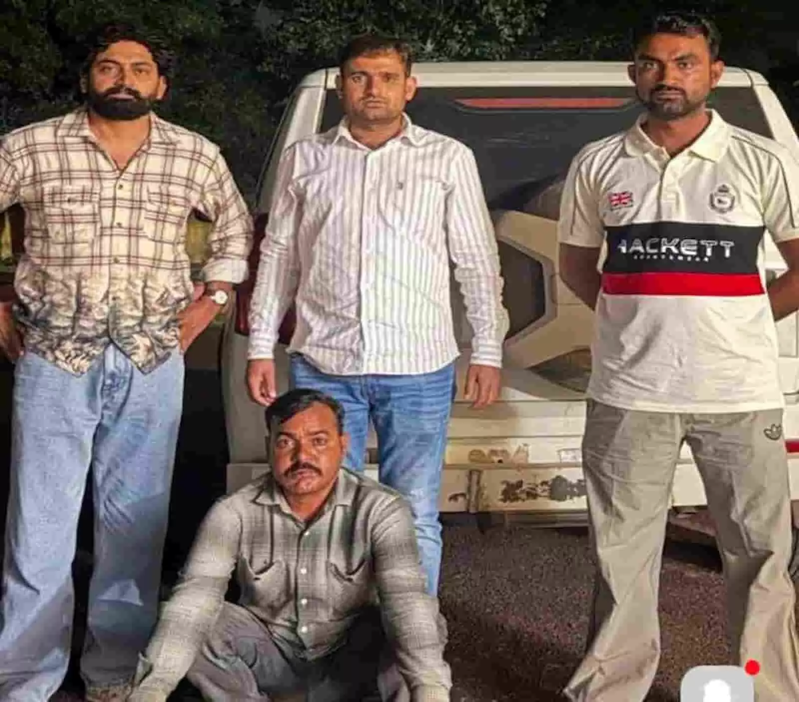 तस्वीर में दिख रहे चार लोगों में से एक व्यक्ति जमीन पर बैठा है और तीन अन्य उसके पीछे खड़े हैं। तस्वीर में दिख रहे चार लोगों में से एक व्यक्ति जमीन पर बैठा है और तीन अन्य उसके पीछे खड़े हैं।