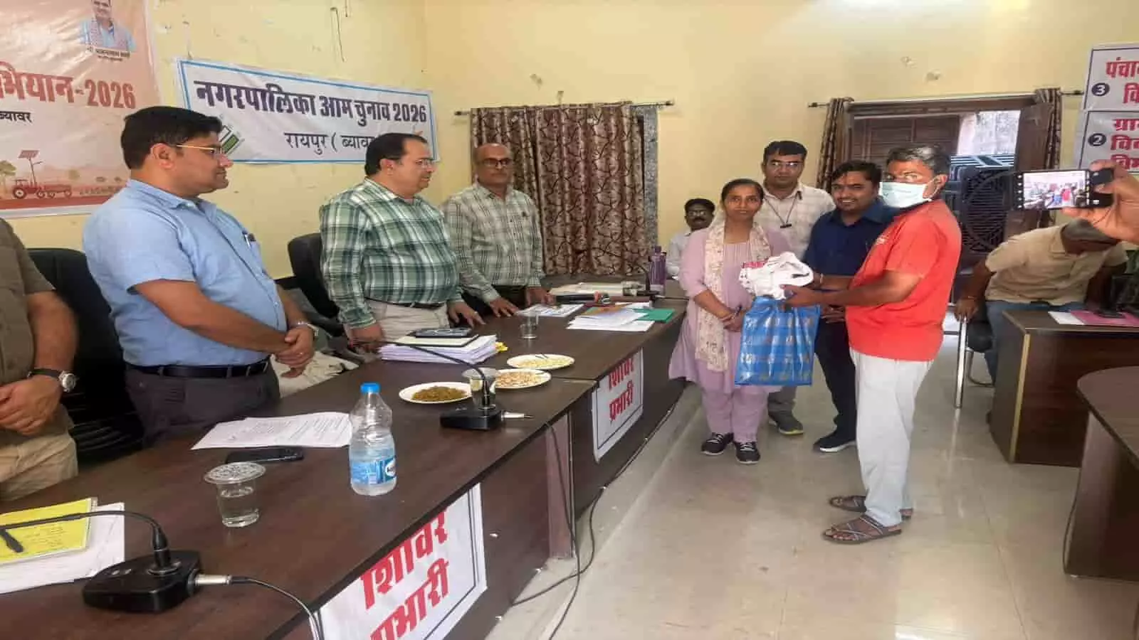तस्वीर में दिख रही एक मेज के पास खड़े अधिकारी एक ग्रामीण व्यक्ति को थैला और आवश्यक सामग्री प्रदान कर रहे हैं। तस्वीर में दिख रही एक मेज के पास खड़े अधिकारी एक ग्रामीण व्यक्ति को थैला और आवश्यक सामग्री प्रदान कर रहे हैं।