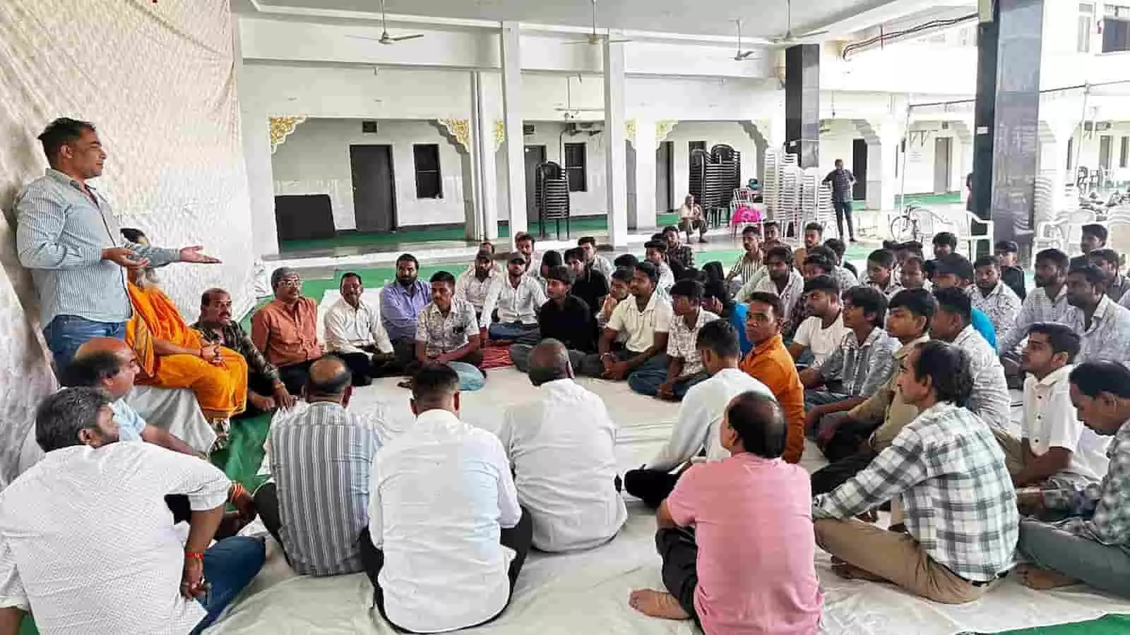 तस्वीर में एक बड़े हॉल में फर्श पर बैठकर सभा को संबोधित करते व्यक्ति को सुनते लोग visible हैं। तस्वीर में एक बड़े हॉल में फर्श पर बैठकर सभा को संबोधित करते व्यक्ति को सुनते लोग visible हैं।