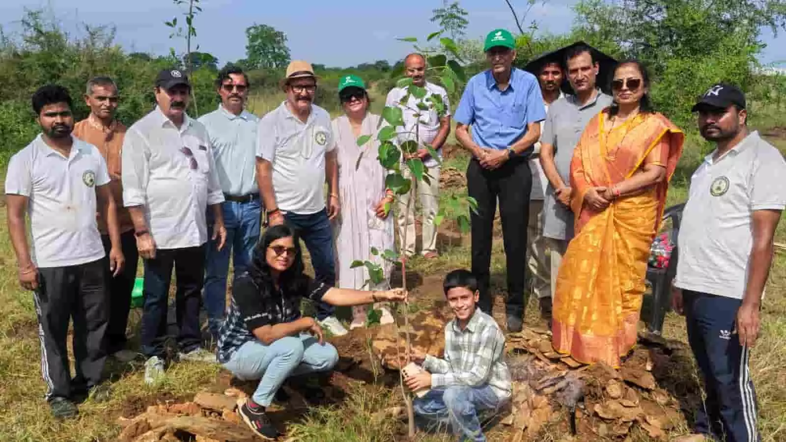 तस्वीर में दिख रहे लोग खुले क्षेत्र में एक छोटे पौधे के साथ खड़े हैं। तस्वीर में दिख रहे लोग खुले क्षेत्र में एक छोटे पौधे के साथ खड़े हैं।