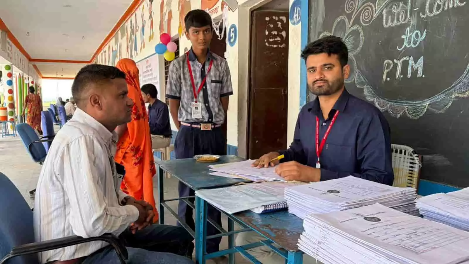 तस्वीर में एक शिक्षक मेज पर बैठकर दस्तावेजों की जांच कर रहे हैं और उनके सामने एक अभिभावक बैठे हैं। तस्वीर में एक शिक्षक मेज पर बैठकर दस्तावेजों की जांच कर रहे हैं और उनके सामने एक अभिभावक बैठे हैं।