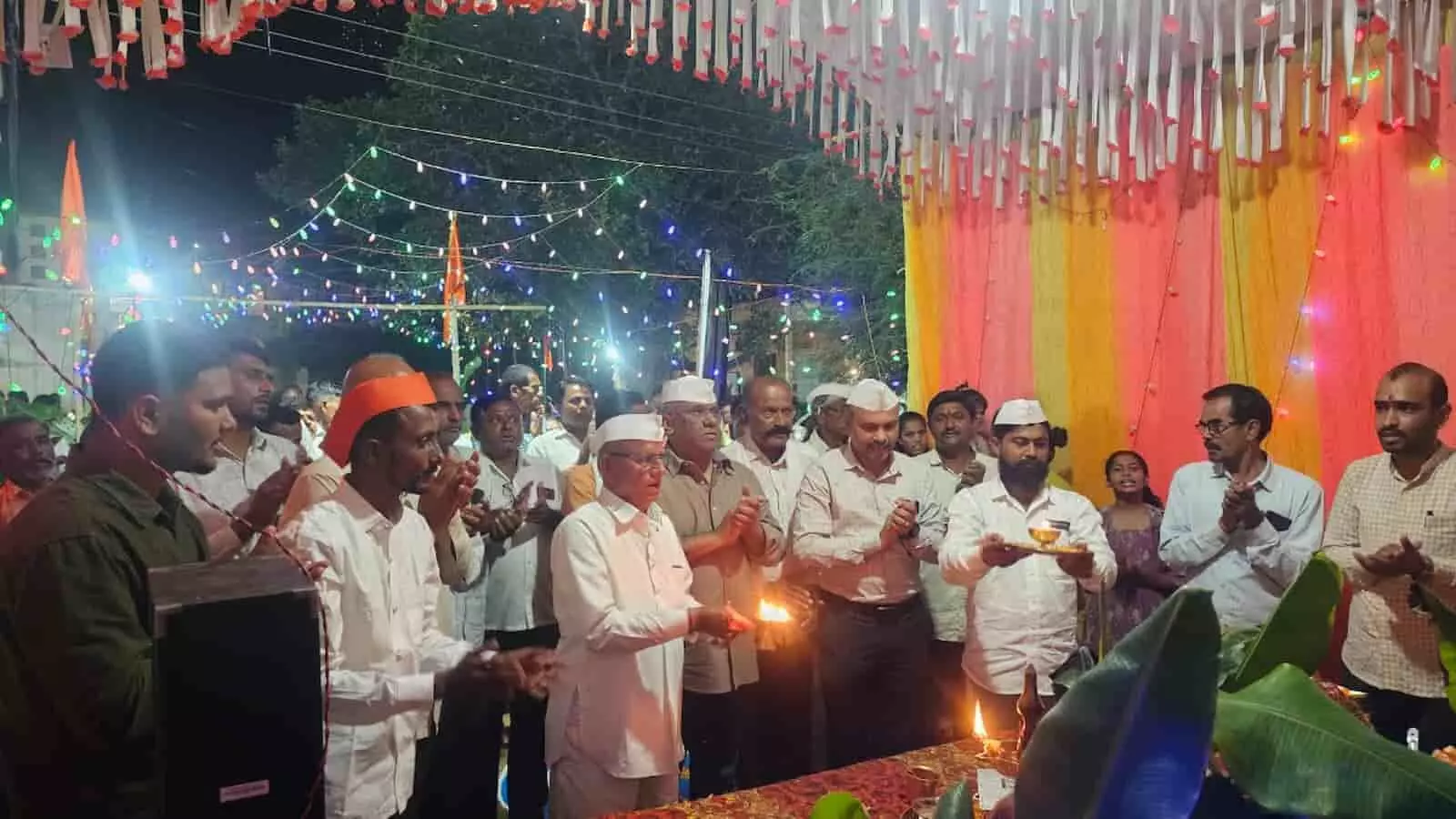 तस्वीर में रोशनी से सजे पंडाल के नीचे भगवान गणेश की आरती करते हुए और हाथ जोड़े खड़े ग्रामीण दिखाई दे रहे हैं। तस्वीर में रोशनी से सजे पंडाल के नीचे भगवान गणेश की आरती करते हुए और हाथ जोड़े खड़े ग्रामीण दिखाई दे रहे हैं।