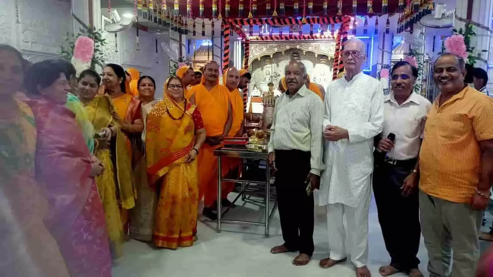 तस्वीर में बाएं से दाएं पारंपरिक वेशभूषा में खड़ी महिलाएं, पीले वस्त्रों में व्यक्ति और सफेद तथा रंगीन कपड़ों में खड़े पुरुष मंदिर परिसर में दिखाई दे रहे हैं। तस्वीर में बाएं से दाएं पारंपरिक वेशभूषा में खड़ी महिलाएं, पीले वस्त्रों में व्यक्ति और सफेद तथा रंगीन कपड़ों में खड़े पुरुष मंदिर परिसर में दिखाई दे रहे हैं।