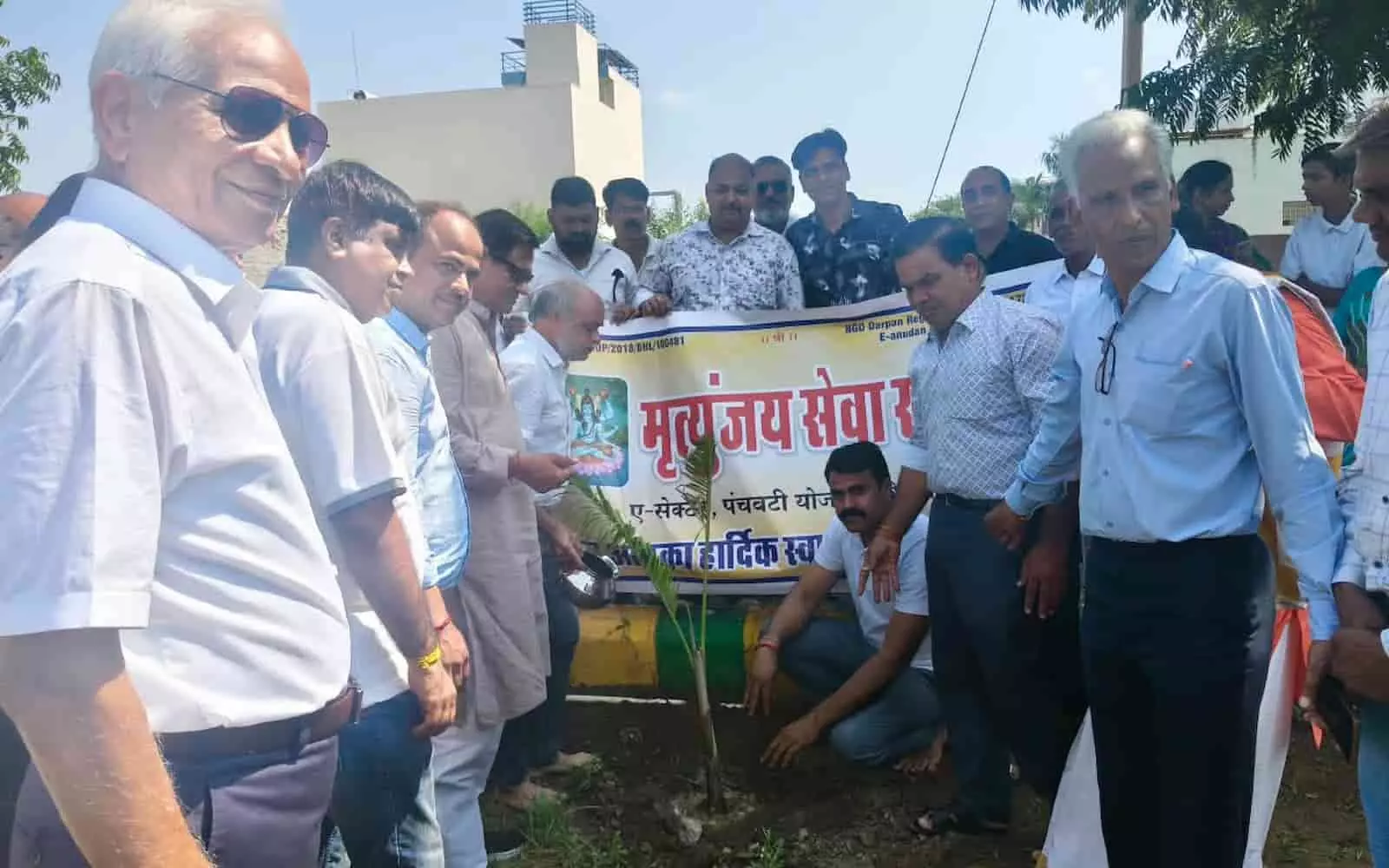 तस्वीर में बाएं से दाएं खड़े हुए कई लोग पौधे के पास और बैनर के पीछे दिखाई दे रहे हैं। तस्वीर में बाएं से दाएं खड़े हुए कई लोग पौधे के पास और बैनर के पीछे दिखाई दे रहे हैं।
