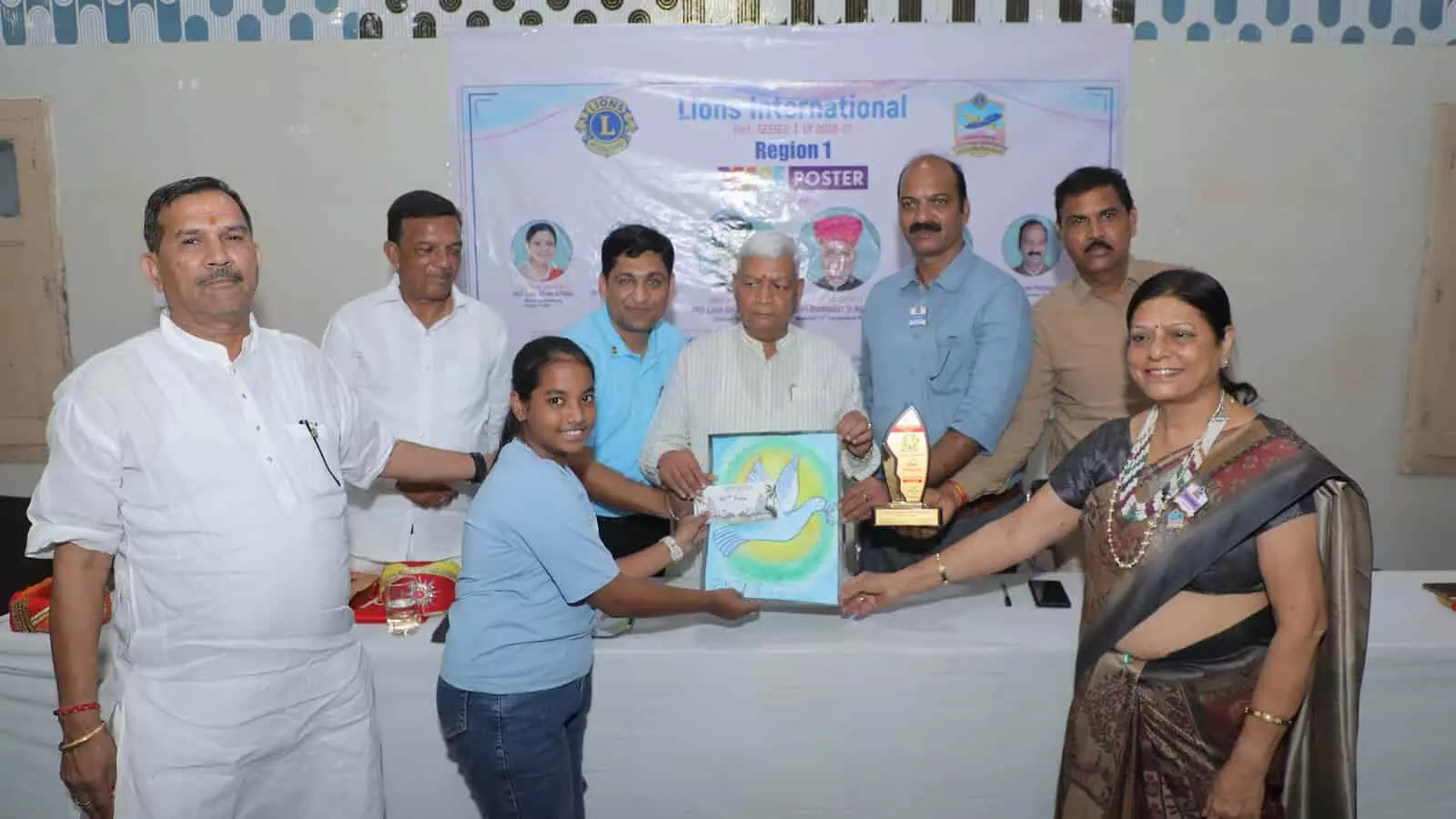 तस्वीर में बाएं से दाएं विभिन्न कपड़ों में खड़े कई लोग और नेता मेज के पास दिखाई दे रहे हैं। तस्वीर में बाएं से दाएं विभिन्न कपड़ों में खड़े कई लोग और नेता मेज के पास दिखाई दे रहे हैं।