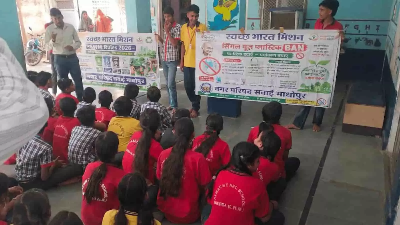 तस्वीर में आगे की तरफ यूनिफॉर्म पहने जमीन पर बैठे विद्यार्थी और पीछे खड़े होकर बैनर पकड़े हुए लोग दिखाई दे रहे हैं। तस्वीर में आगे की तरफ यूनिफॉर्म पहने जमीन पर बैठे विद्यार्थी और पीछे खड़े होकर बैनर पकड़े हुए लोग दिखाई दे रहे हैं।