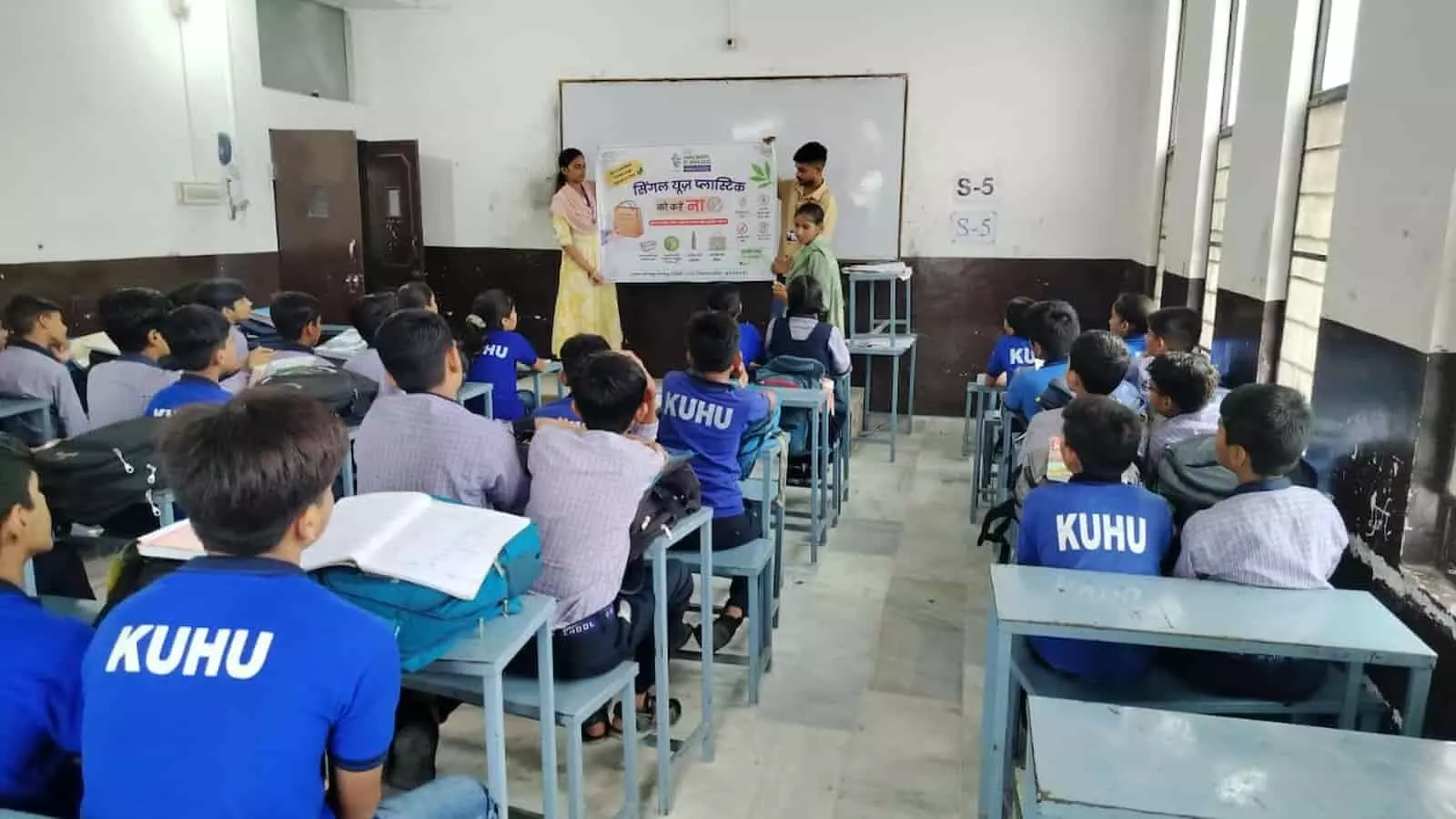तस्वीर में आगे बेंचों पर बैठे हुए यूनिफॉर्म पहने विद्यार्थी और सामने खड़े होकर बैनर दिखाते हुए लोग दिखाई दे रहे हैं। तस्वीर में आगे बेंचों पर बैठे हुए यूनिफॉर्म पहने विद्यार्थी और सामने खड़े होकर बैनर दिखाते हुए लोग दिखाई दे रहे हैं।