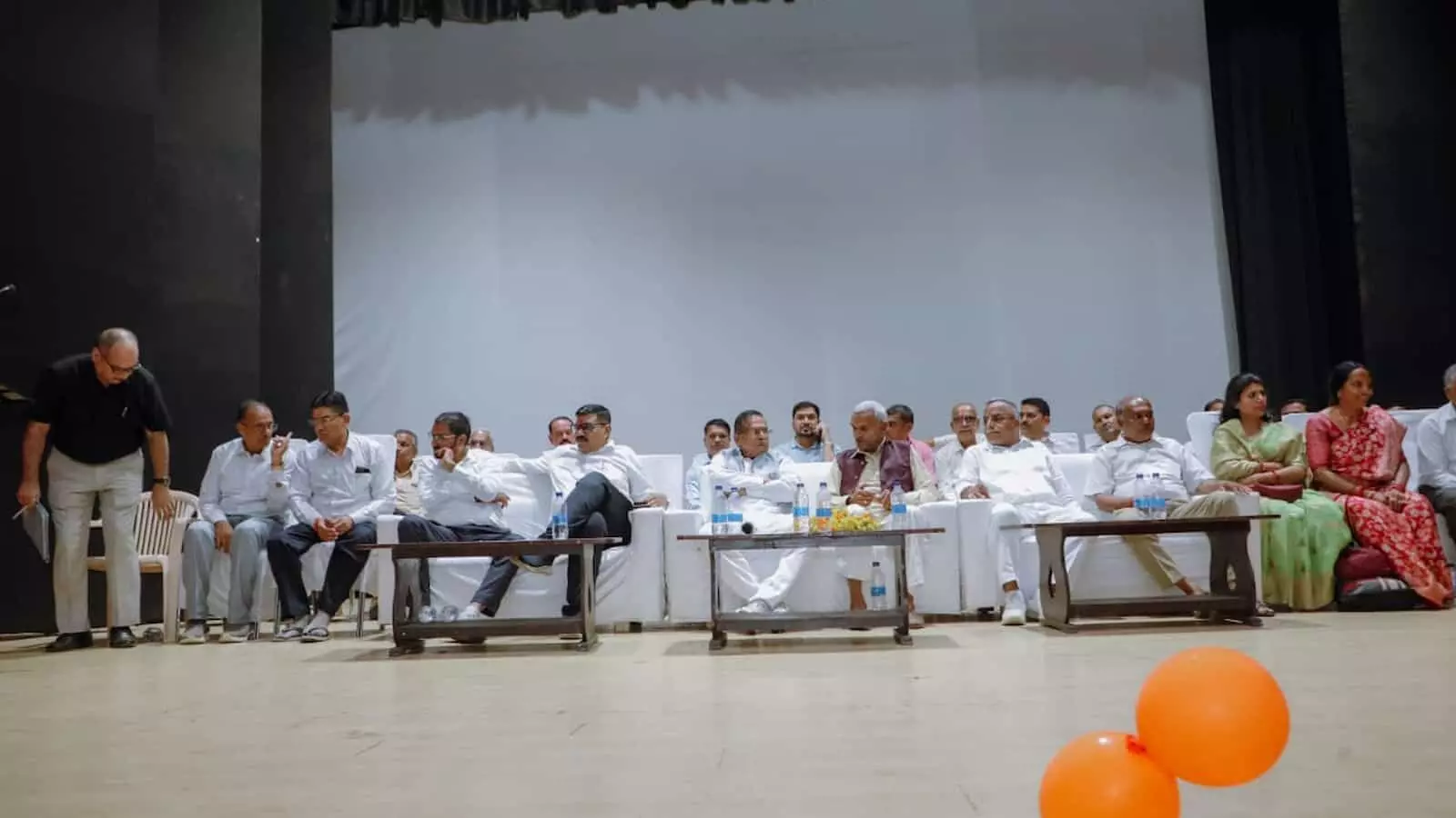 तस्वीर में ऑडिटोरियम के मंच पर सफेद कुर्सियों पर बैठे हुए कई पुरुष और महिलाएं दिखाई दे रहे हैं। तस्वीर में ऑडिटोरियम के मंच पर सफेद कुर्सियों पर बैठे हुए कई पुरुष और महिलाएं दिखाई दे रहे हैं।