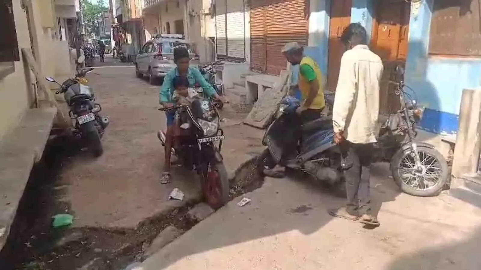 क्षतिग्रस्त नाली के पास गली में खड़े लोग और वाहन चौमहला में। क्षतिग्रस्त नाली के पास गली में खड़े लोग और वाहन चौमहला में।