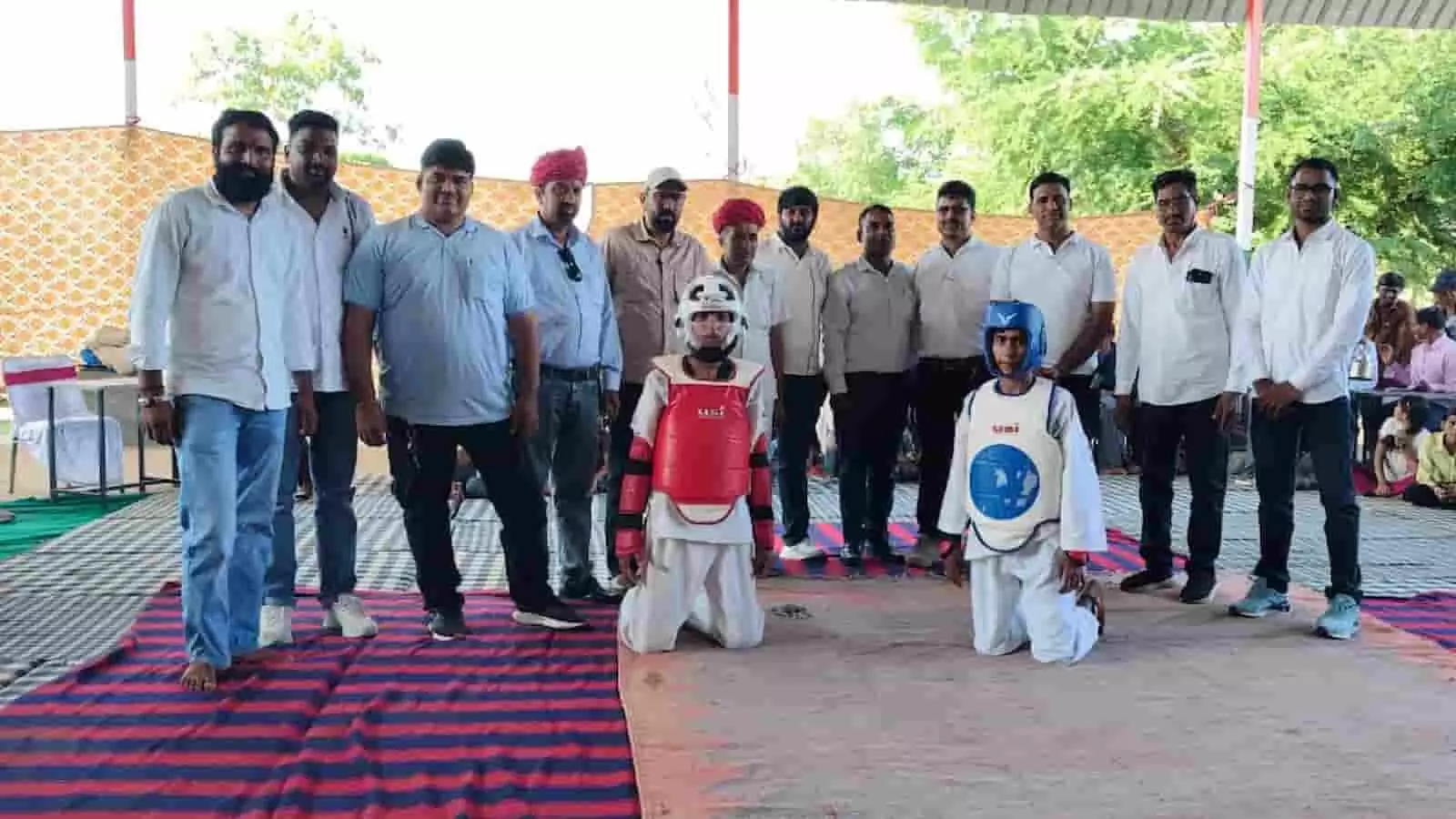 तस्वीर में ताइक्वांडो पोशाक पहने दो खिलाड़ी घुटनों के बल बैठे हैं और पीछे अतिथि खड़े हैं। तस्वीर में ताइक्वांडो पोशाक पहने दो खिलाड़ी घुटनों के बल बैठे हैं और पीछे अतिथि खड़े हैं।