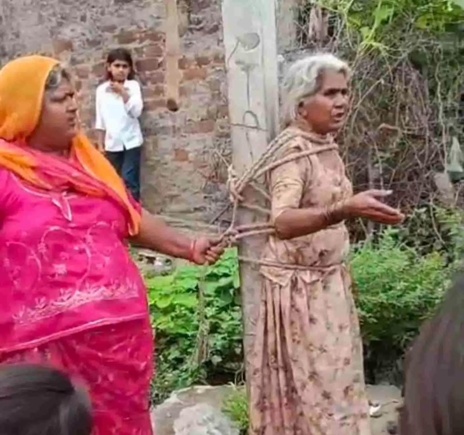 तस्वीर में बाएं ओर खड़ी महिला और दाएं ओर खंभे से रस्सी से बंधी बुजुर्ग महिला दिखाई दे रही हैं। तस्वीर में बाएं ओर खड़ी महिला और दाएं ओर खंभे से रस्सी से बंधी बुजुर्ग महिला दिखाई दे रही हैं।