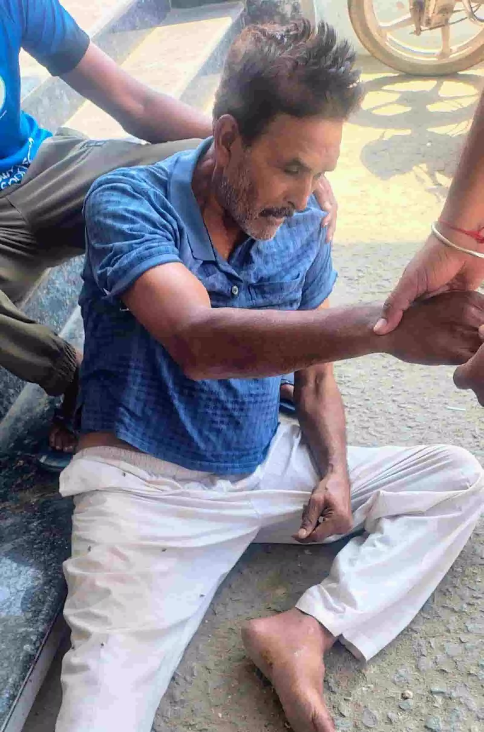 तस्वीर में दिख रही जगह पर एक व्यक्ति सड़क किनारे बैठा है और अन्य लोग उसकी नाड़ी जांच रहे हैं। तस्वीर में दिख रही जगह पर एक व्यक्ति सड़क किनारे बैठा है और अन्य लोग उसकी नाड़ी जांच रहे हैं।