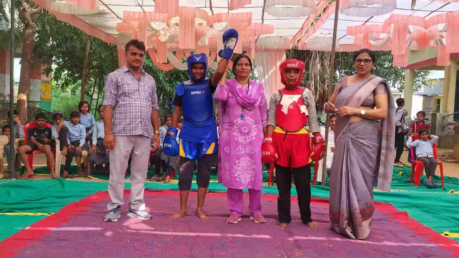 तस्वीर में बाएं से दाएं एक पुरुष, नीली पोशाक पहने खिलाड़ी, बीच में महिला, लाल पोशाक पहने खिलाड़ी और दाईं ओर साड़ी पहनी महिला खड़ी हैं. तस्वीर में बाएं से दाएं एक पुरुष, नीली पोशाक पहने खिलाड़ी, बीच में महिला, लाल पोशाक पहने खिलाड़ी और दाईं ओर साड़ी पहनी महिला खड़ी हैं.