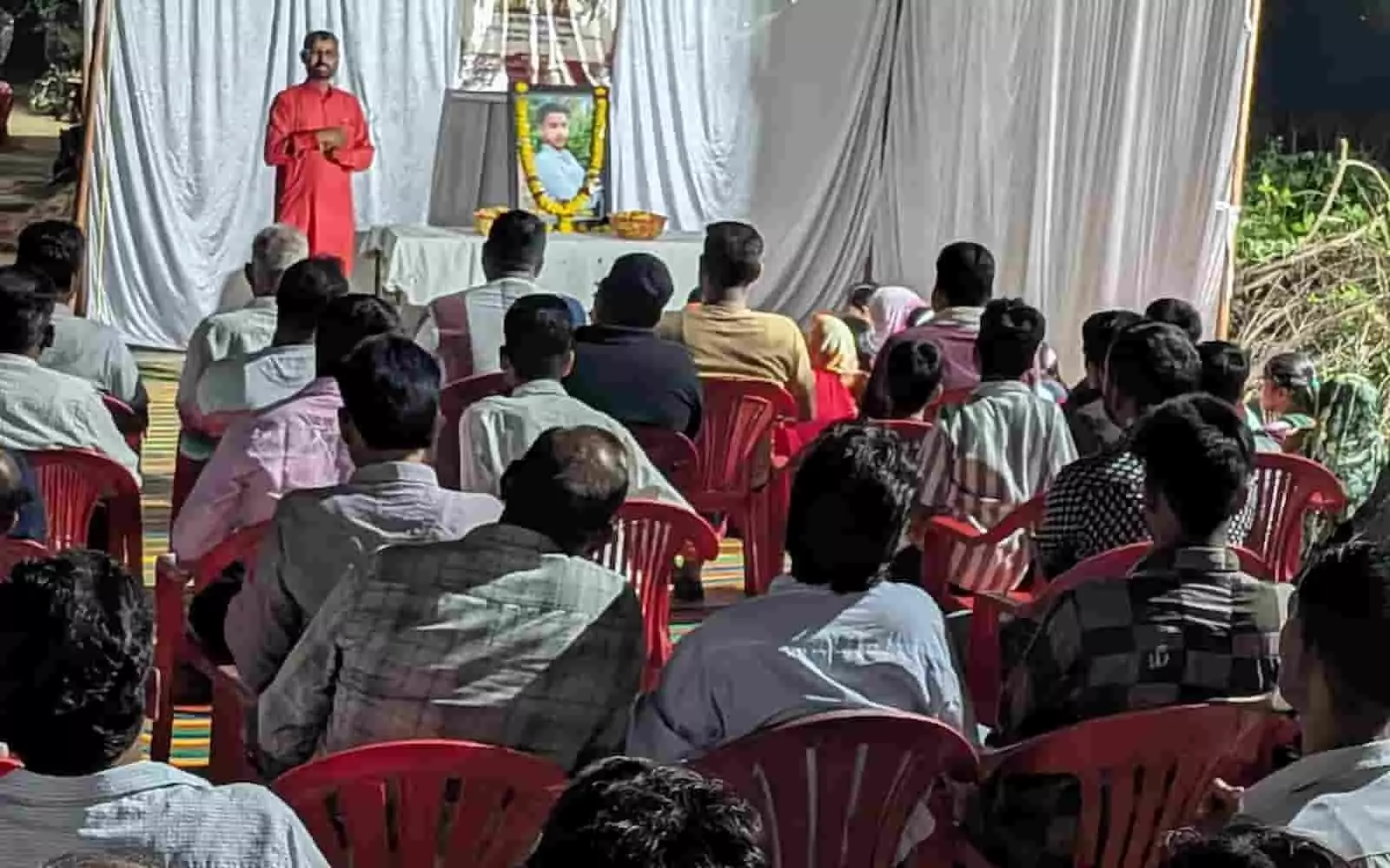 तस्वीर में दिख रही सभा में एक व्यक्ति मंच पर बोल रहा है और सामने कुर्सियों पर लोग बैठे हैं। तस्वीर में दिख रही सभा में एक व्यक्ति मंच पर बोल रहा है और सामने कुर्सियों पर लोग बैठे हैं।