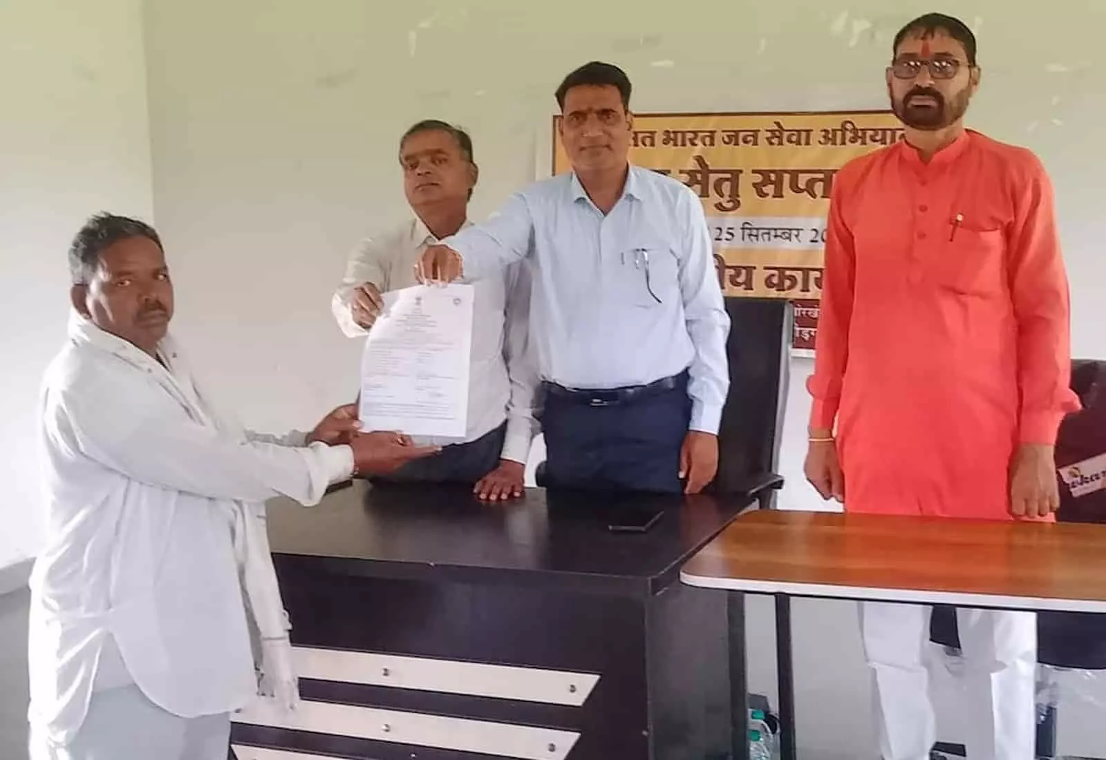 तस्वीर में बाएं खड़े अधिकारी एक व्यक्ति को दस्तावेज सौंप रहे हैं और अन्य अधिकारी पास खड़े हैं। तस्वीर में बाएं खड़े अधिकारी एक व्यक्ति को दस्तावेज सौंप रहे हैं और अन्य अधिकारी पास खड़े हैं।