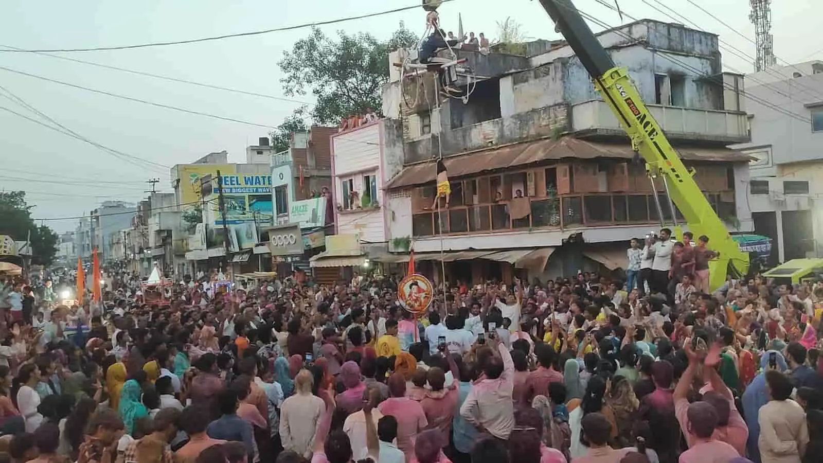 तस्वीर में एक सड़क पर शोभायात्रा के दौरान लोगों की भीड़ और ऊपर क्रेन से बंधे करतब दिखाते हुए लोग दिख रहे हैं। तस्वीर में एक सड़क पर शोभायात्रा के दौरान लोगों की भीड़ और ऊपर क्रेन से बंधे करतब दिखाते हुए लोग दिख रहे हैं।
