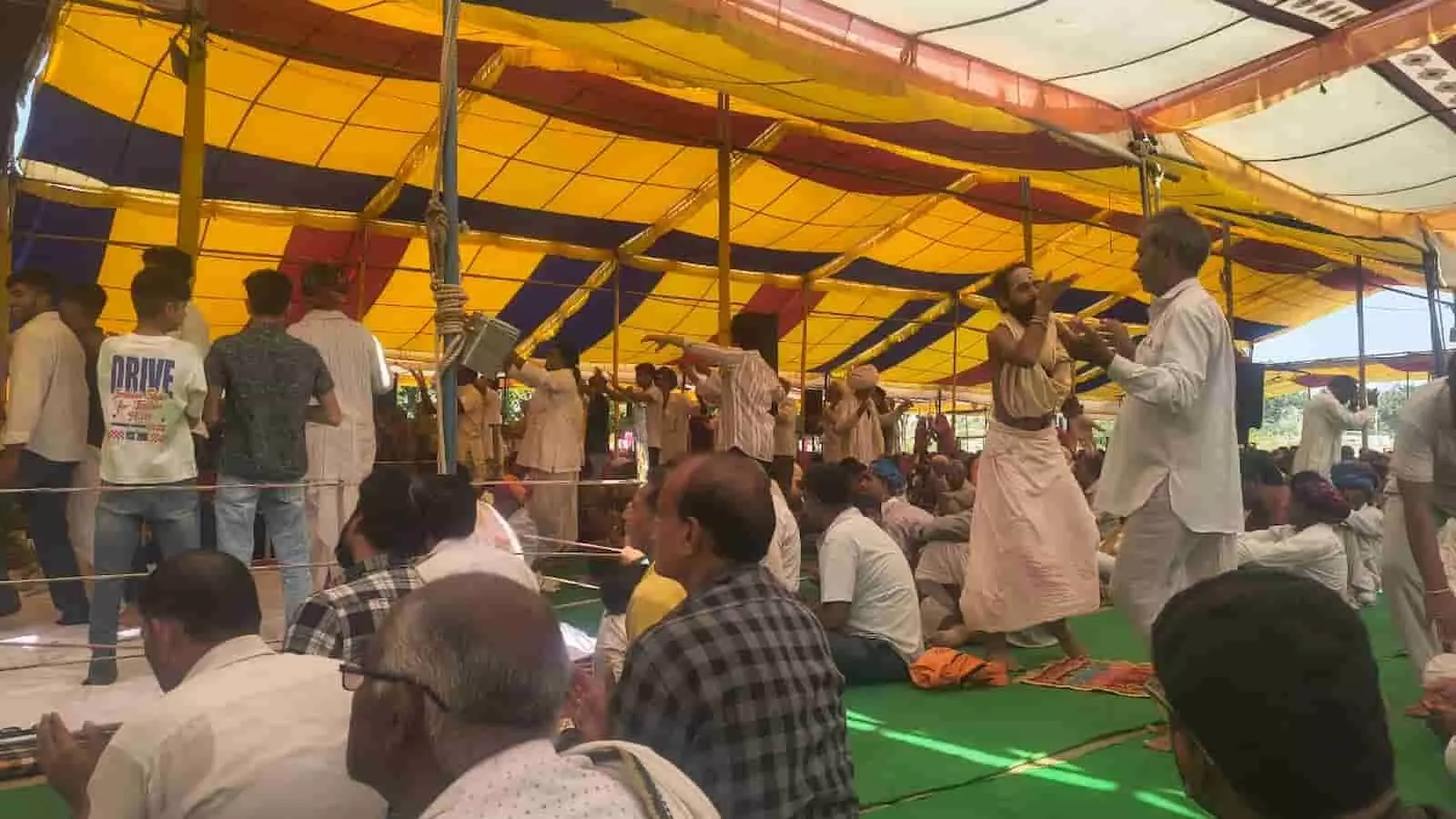 तस्वीर में पंडाल के नीचे बैठे लोग और सामने खड़े होकर ताली बजाते श्रद्धालु दिख रहे हैं। तस्वीर में पंडाल के नीचे बैठे लोग और सामने खड़े होकर ताली बजाते श्रद्धालु दिख रहे हैं।