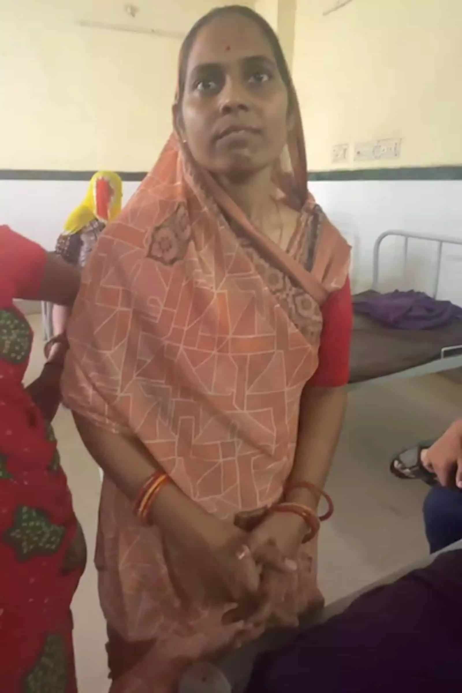 अस्पताल के वार्ड में अन्य लोगों के पास खड़ी गुलाबी साड़ी पहनी एक महिला अस्पताल के वार्ड में अन्य लोगों के पास खड़ी गुलाबी साड़ी पहनी एक महिला