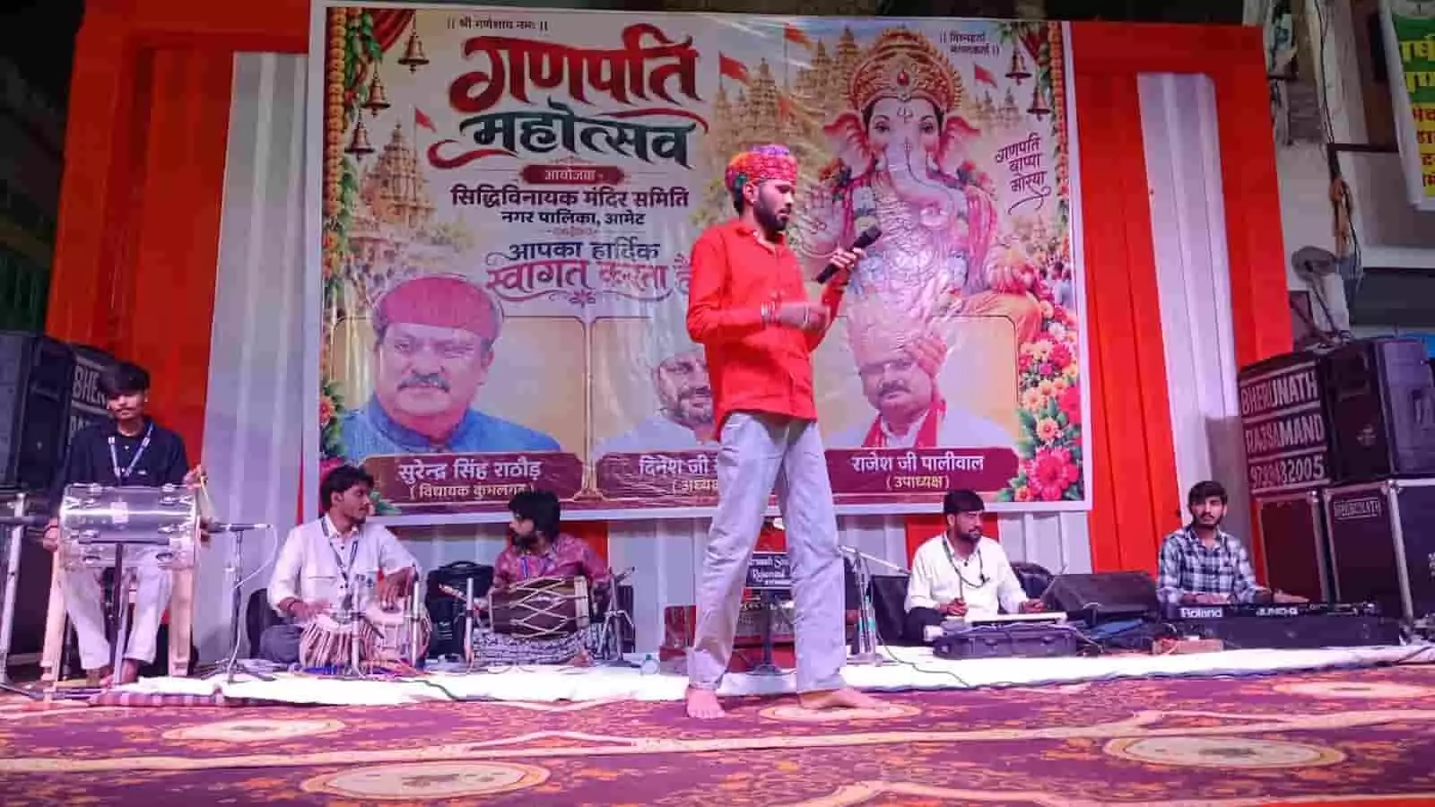 तस्वीर में लाल शर्ट पहने गायक मंच पर हाथ में माइक लिए खड़े हैं और वाद्ययंत्र वादक साथ बैठे हैं। तस्वीर में लाल शर्ट पहने गायक मंच पर हाथ में माइक लिए खड़े हैं और वाद्ययंत्र वादक साथ बैठे हैं।