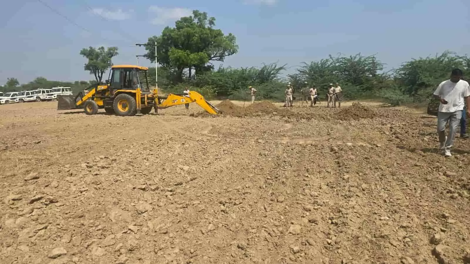 तस्वीर में खुले मैदान में खड़े सुरक्षाकर्मियों और पुलिस वाहनों के बीच जमीन की खुदाई करती पीली जेसीबी मशीन दिखाई दे रही है। तस्वीर में खुले मैदान में खड़े सुरक्षाकर्मियों और पुलिस वाहनों के बीच जमीन की खुदाई करती पीली जेसीबी मशीन दिखाई दे रही है।