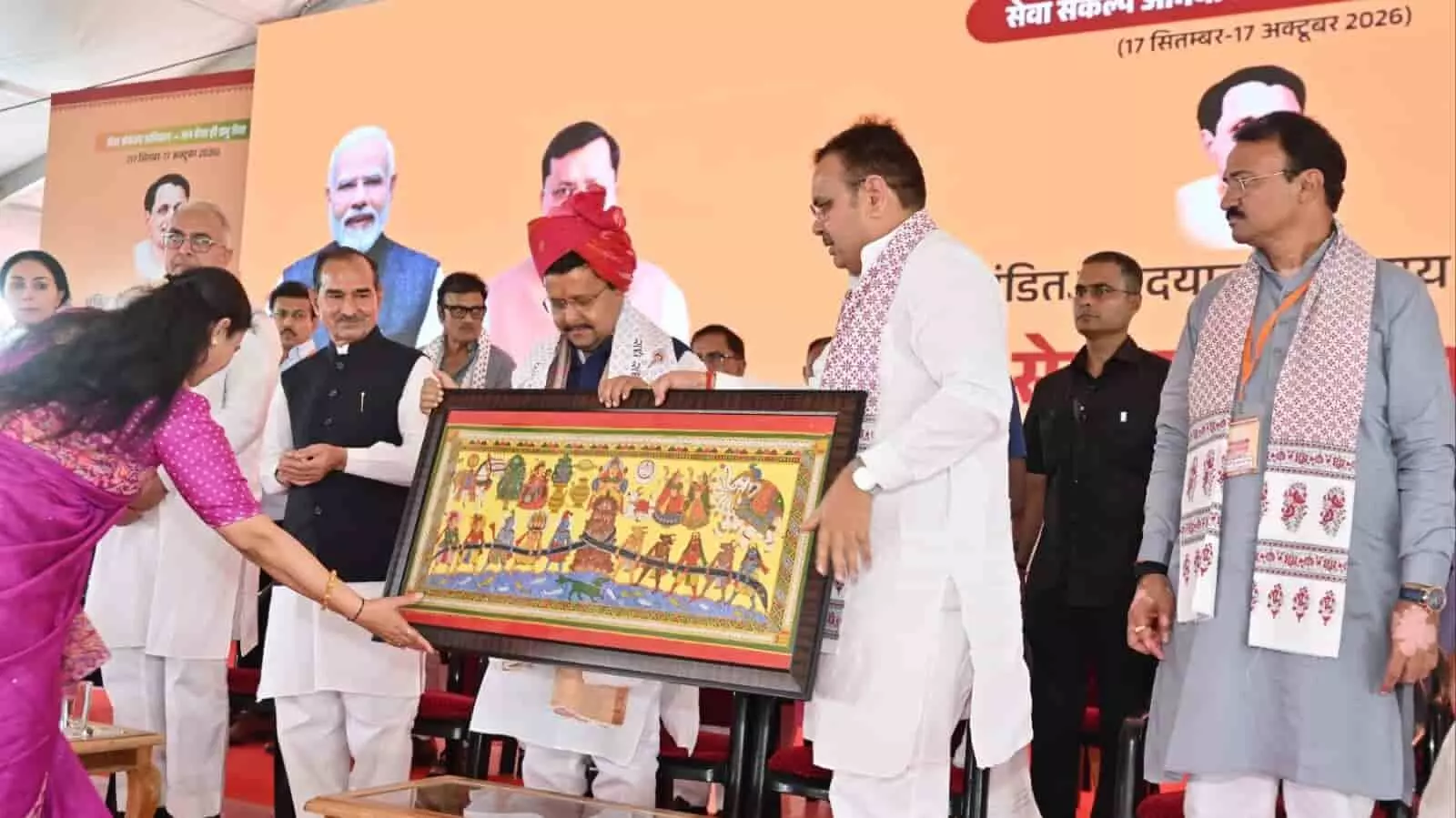 तस्वीर में पारंपरिक साफा पहने हुए नेताओं को मंच पर एक बड़ी तख्ती या फ्रेम भेंट करते हुए लोग दिखाई दे रहे हैं। तस्वीर में पारंपरिक साफा पहने हुए नेताओं को मंच पर एक बड़ी तख्ती या फ्रेम भेंट करते हुए लोग दिखाई दे रहे हैं।