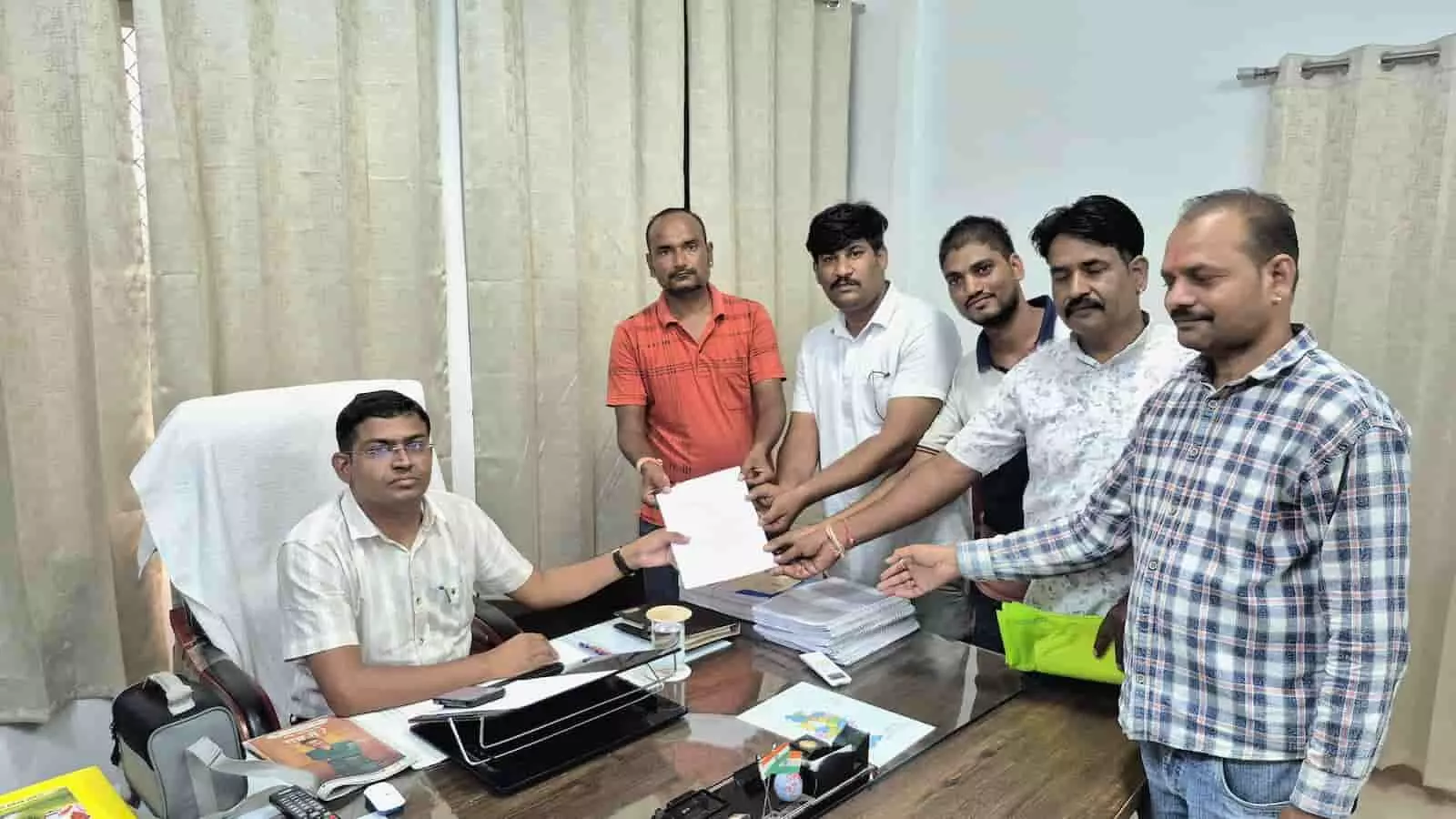 तस्वीर में एक अधिकारी अपनी मेज पर बैठे हैं और ग्रामीण उन्हें ज्ञापन सौंप रहे हैं। तस्वीर में एक अधिकारी अपनी मेज पर बैठे हैं और ग्रामीण उन्हें ज्ञापन सौंप रहे हैं।