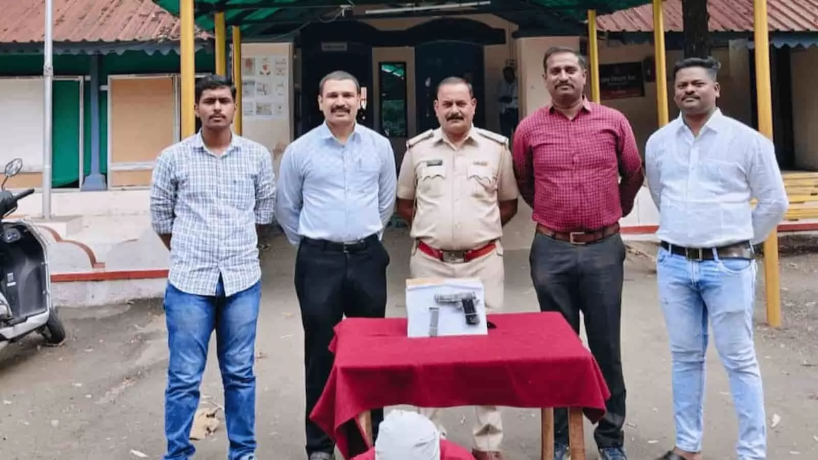 तस्वीर में पुलिस स्टेशन के बाहर मेज पर रखी पिस्तौल के पास नीचे बैठे संदिग्ध युवक और खड़े पुलिस अधिकारियों का समूह दिखाई दे रहा है। तस्वीर में पुलिस स्टेशन के बाहर मेज पर रखी पिस्तौल के पास नीचे बैठे संदिग्ध युवक और खड़े पुलिस अधिकारियों का समूह दिखाई दे रहा है।