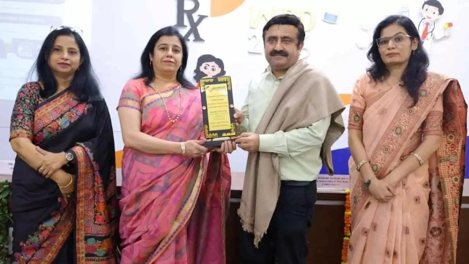 तस्वीर में बाएं से दाएं चार लोग खड़े हैं, जो मंच पर एक स्मृति चिह्न थामे हुए हैं। तस्वीर में बाएं से दाएं चार लोग खड़े हैं, जो मंच पर एक स्मृति चिह्न थामे हुए हैं।