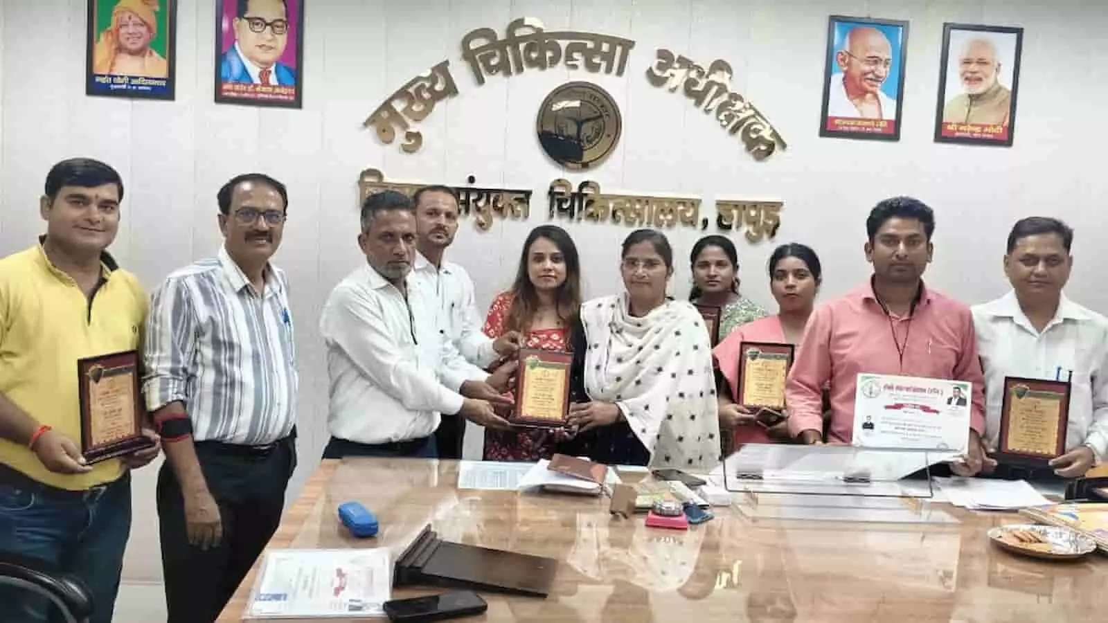 तस्वीर में बाएं से दाएं कई डॉक्टर और स्वास्थ्यकर्मी कार्यालय के अंदर हाथ में शील्ड थामे हुए खड़े हैं। तस्वीर में बाएं से दाएं कई डॉक्टर और स्वास्थ्यकर्मी कार्यालय के अंदर हाथ में शील्ड थामे हुए खड़े हैं।