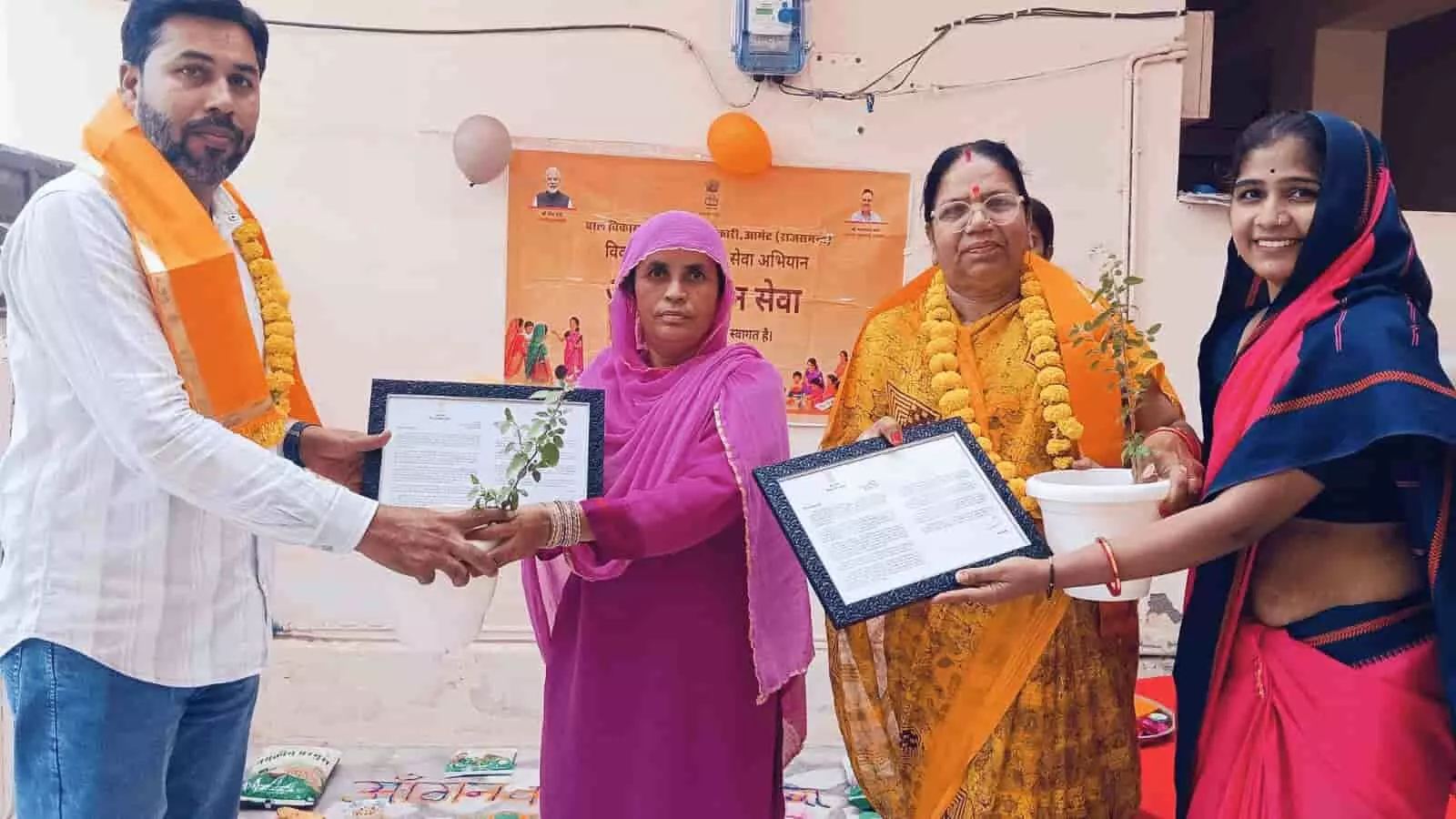 तस्वीर में बाएं से दाएं एक व्यक्ति और कुछ महिलाएं आंगनवाड़ी केंद्र पर खड़े होकर हाथ में पौधे का गमला और प्रमाण पत्र पकड़े हुए हैं। तस्वीर में बाएं से दाएं एक व्यक्ति और कुछ महिलाएं आंगनवाड़ी केंद्र पर खड़े होकर हाथ में पौधे का गमला और प्रमाण पत्र पकड़े हुए हैं।