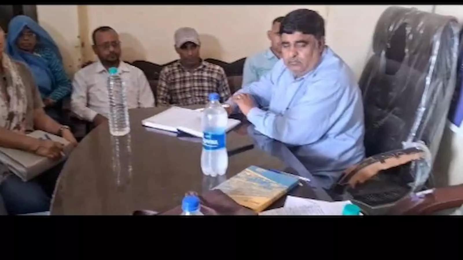 तस्वीर में मेज के पास बैठे अधिकारी और पीछे कुछ लोग चर्चा करते नजर आ रहे हैं। तस्वीर में मेज के पास बैठे अधिकारी और पीछे कुछ लोग चर्चा करते नजर आ रहे हैं।