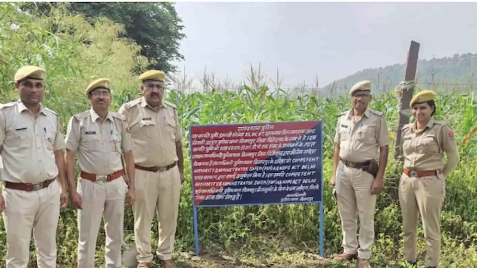तस्वीर में खेत के पास खड़े पुलिसकर्मी और बीच में एक सरकारी सूचना का बोर्ड दिखाई दे रहा है। तस्वीर में खेत के पास खड़े पुलिसकर्मी और बीच में एक सरकारी सूचना का बोर्ड दिखाई दे रहा है।