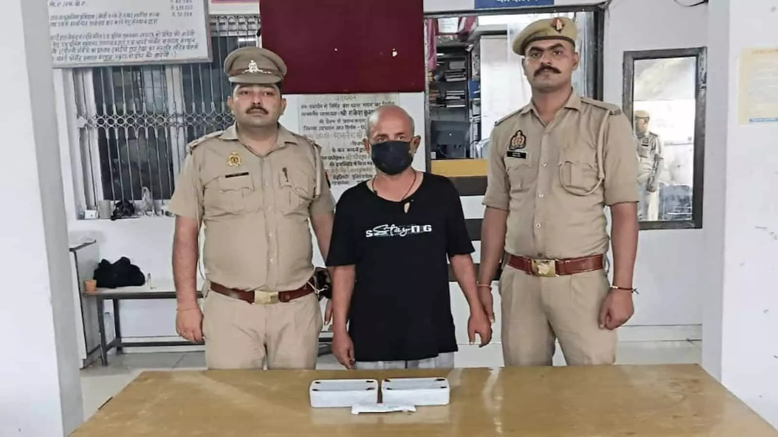 तस्वीर में बीच में खड़े आरोपी के दोनों तरफ खाकी वर्दी पहने दो पुलिसकर्मी खड़े दिखाई दे रहे हैं। तस्वीर में बीच में खड़े आरोपी के दोनों तरफ खाकी वर्दी पहने दो पुलिसकर्मी खड़े दिखाई दे रहे हैं।