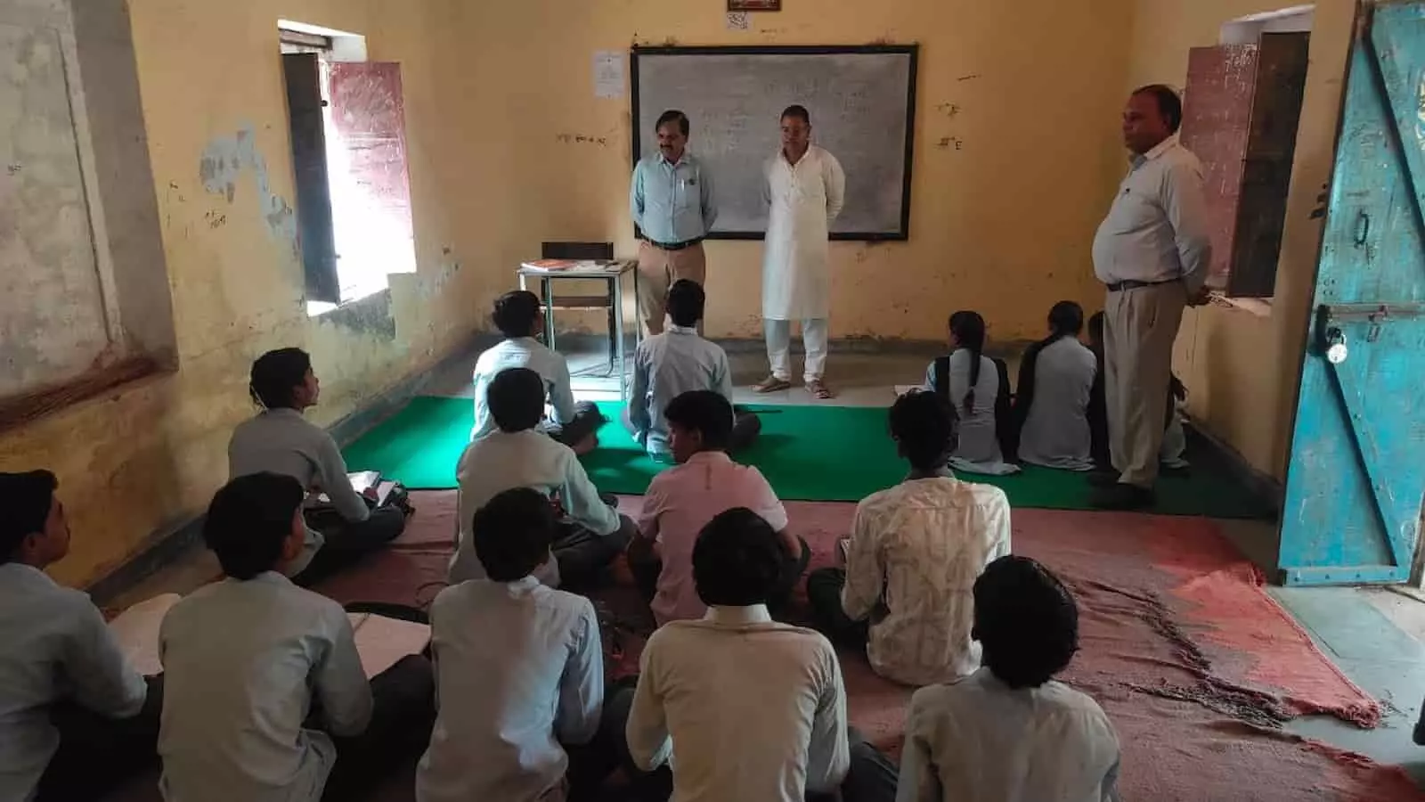 सरकारी स्कूल के कमरे में खड़े अधिकारी और जमीन पर बैठे छात्र-छात्राएं। सरकारी स्कूल के कमरे में खड़े अधिकारी और जमीन पर बैठे छात्र-छात्राएं।