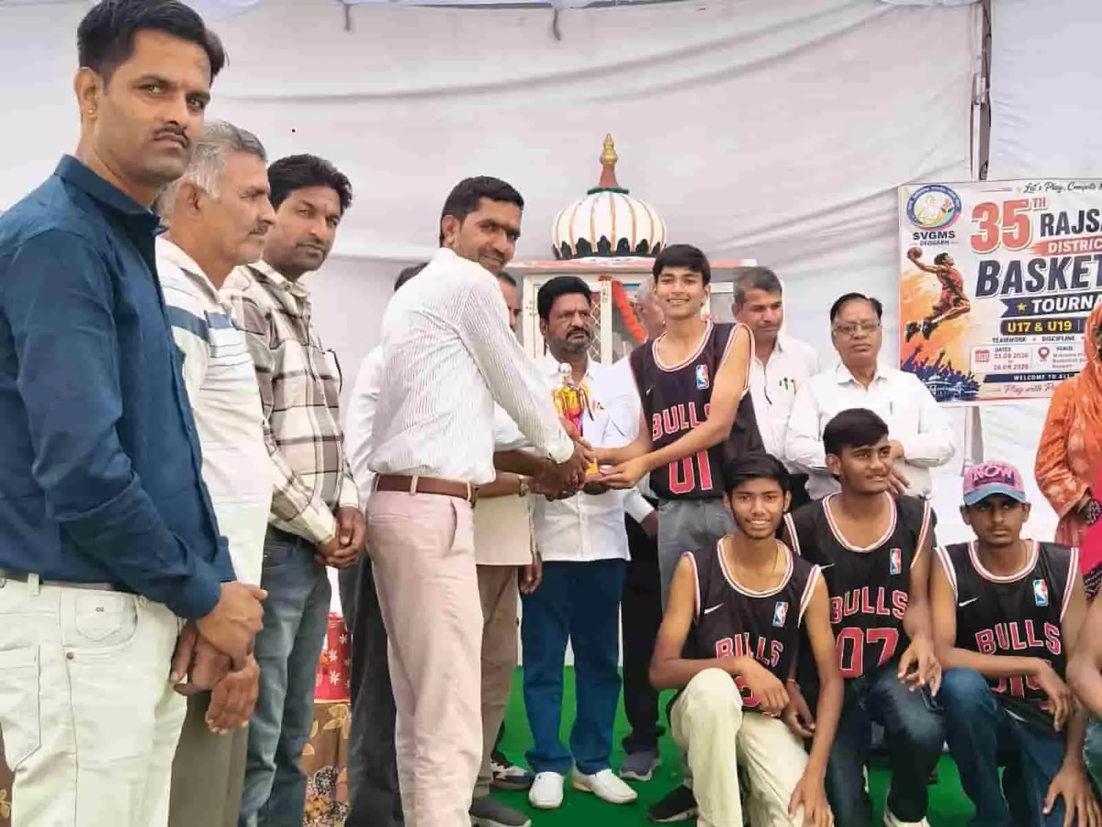 बास्केटबॉल टीम के छात्र और शिक्षक मंच पर ट्रॉफी लेते हुए दिखाई दे रहे हैं। बास्केटबॉल टीम के छात्र और शिक्षक मंच पर ट्रॉफी लेते हुए दिखाई दे रहे हैं।