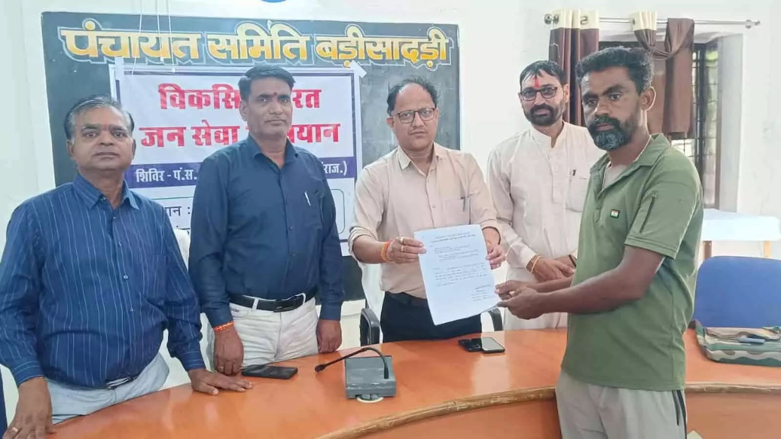 तस्वीर में बाएं से दाएं खड़े अधिकारी कार्यालय में एक व्यक्ति को दस्तावेज प्रदान कर रहे हैं। तस्वीर में बाएं से दाएं खड़े अधिकारी कार्यालय में एक व्यक्ति को दस्तावेज प्रदान कर रहे हैं।